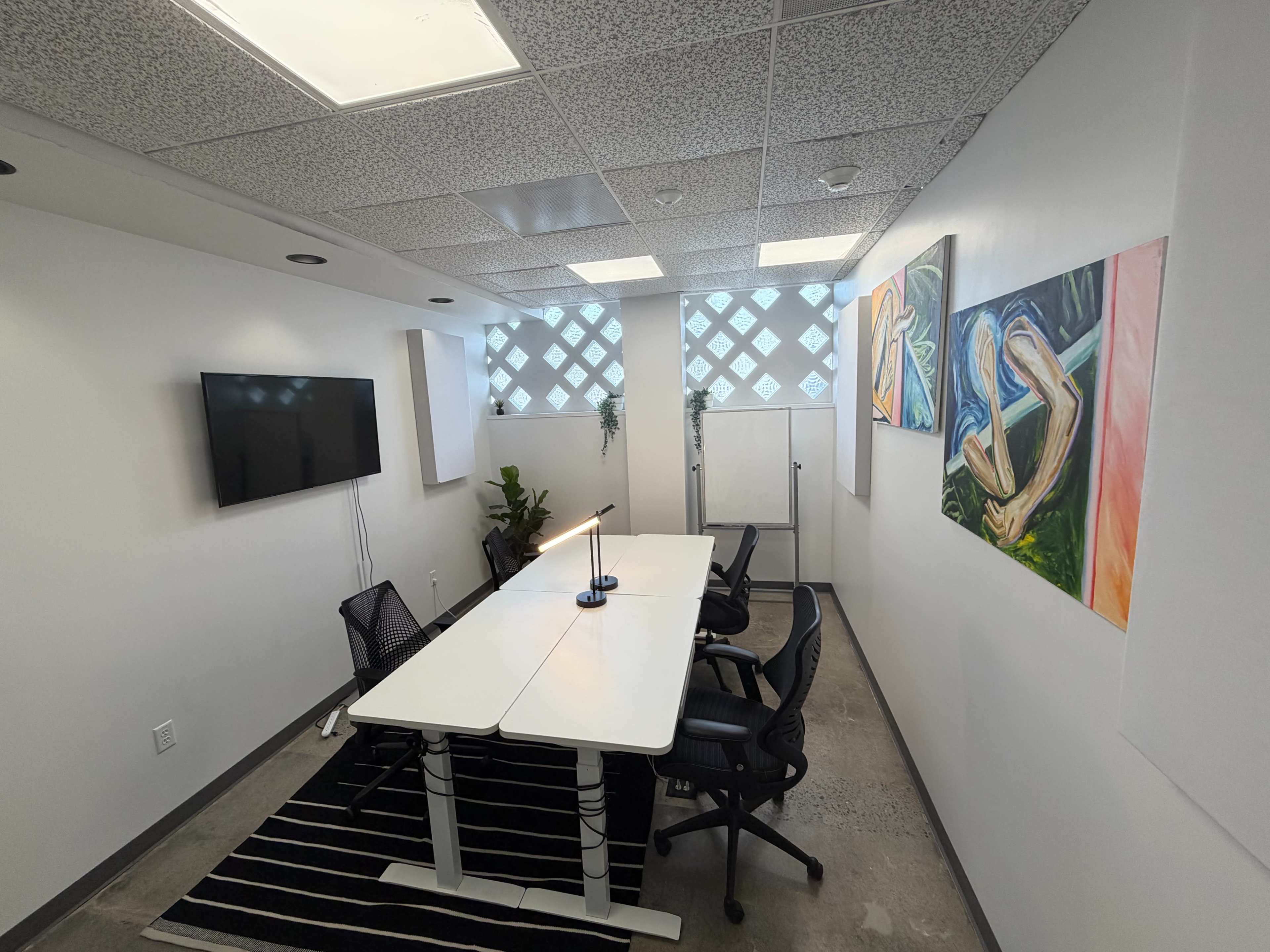 The image shows a conference room featuring a white table, black chairs, a TV on the wall, a whiteboard, and colorful artwork on the walls.