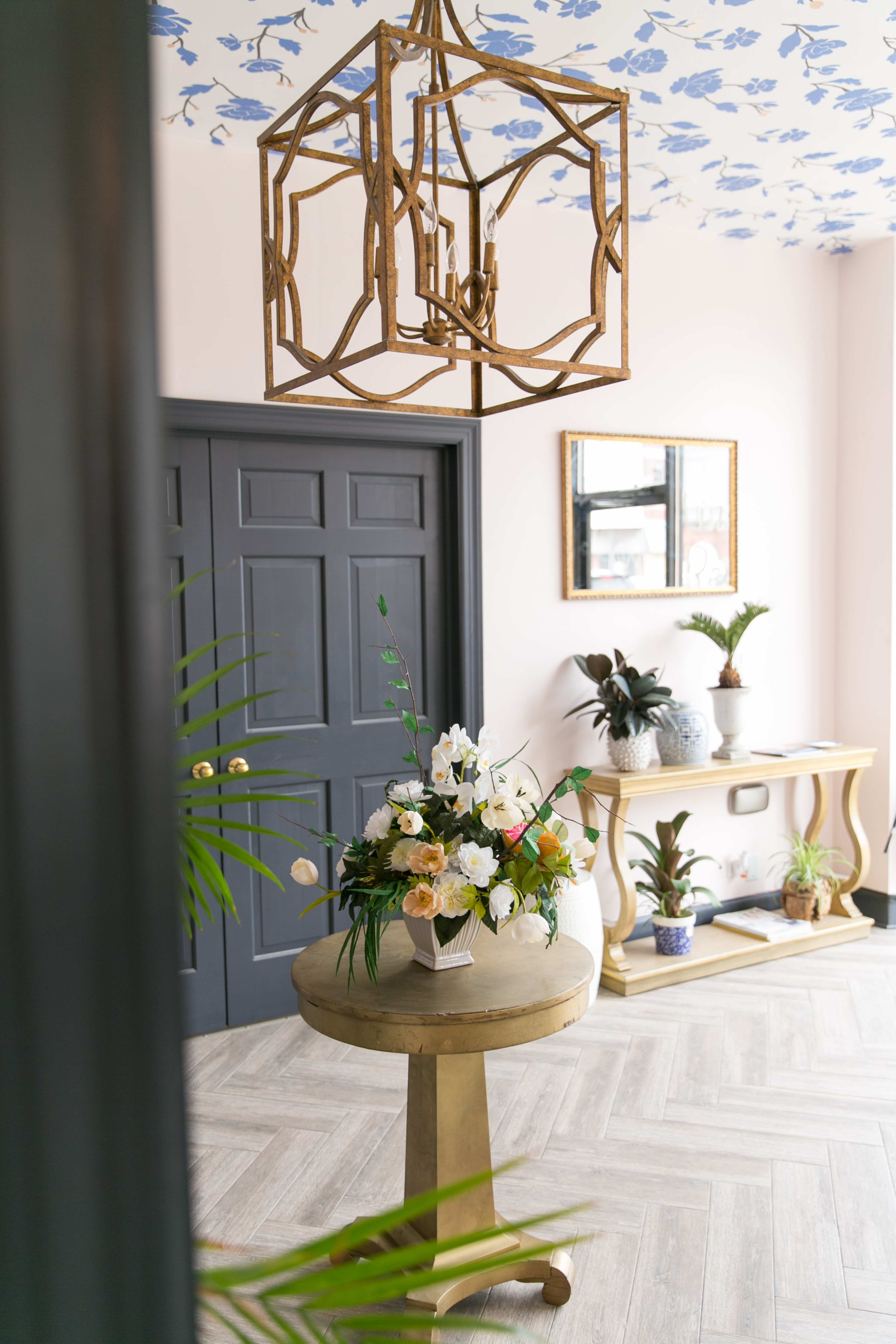 A bright entryway features a decorative chandelier, a round table with a floral arrangement, and plants against a floral wallpaper and dark grey door.