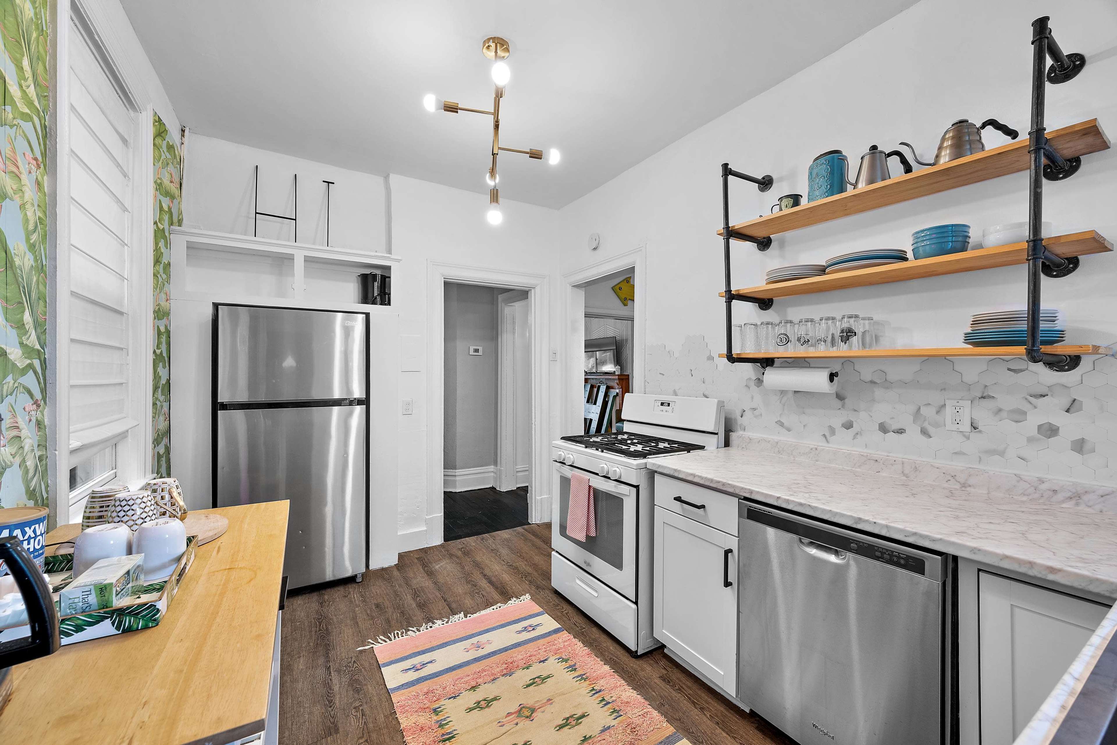 The kitchen features stainless steel appliances, open shelving with dishware, and a patterned rug on the wooden floor.