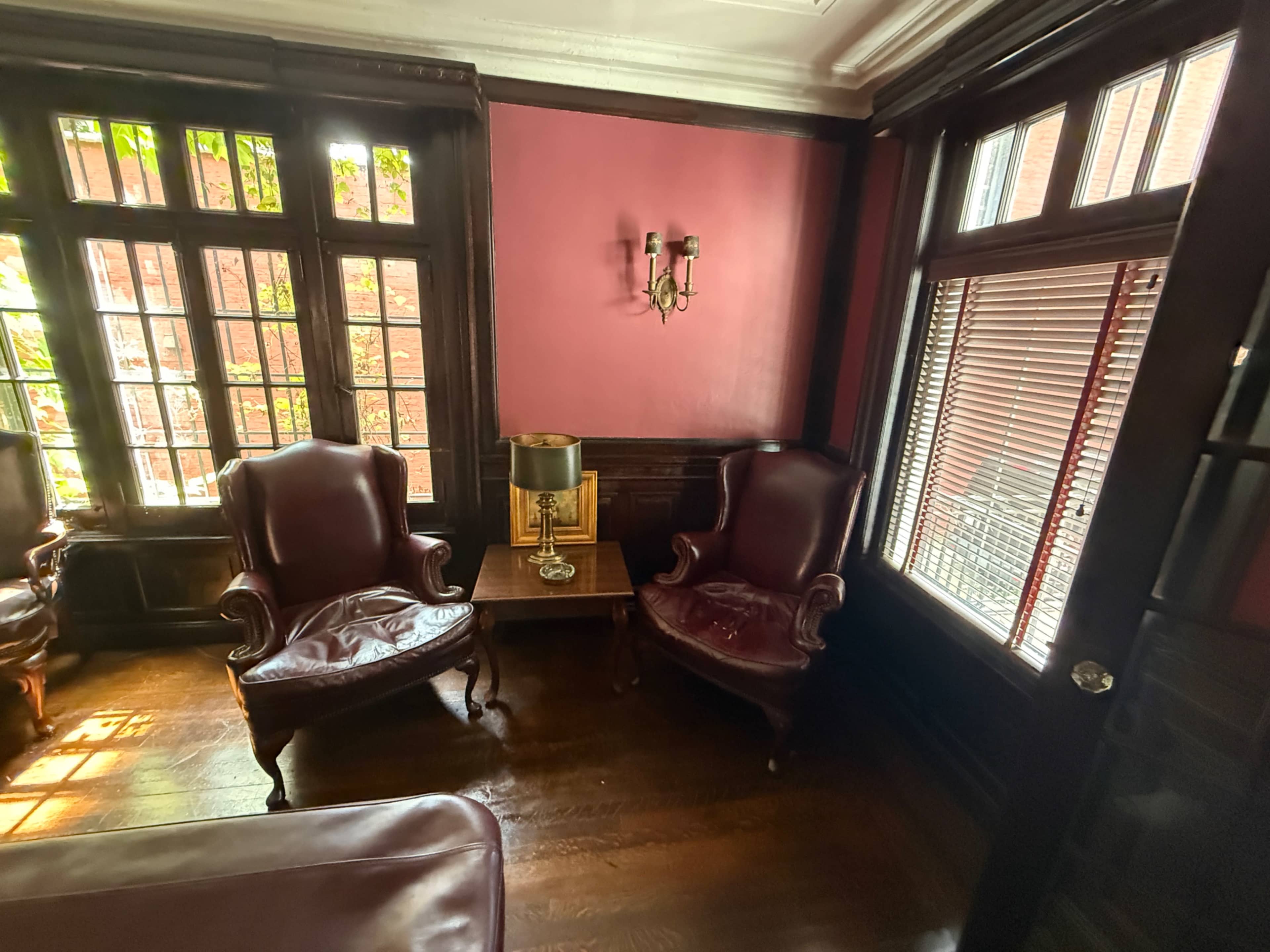 The image shows a cozy room with two burgundy armchairs, a small side table with a lamp, and large windows allowing natural light to enter.