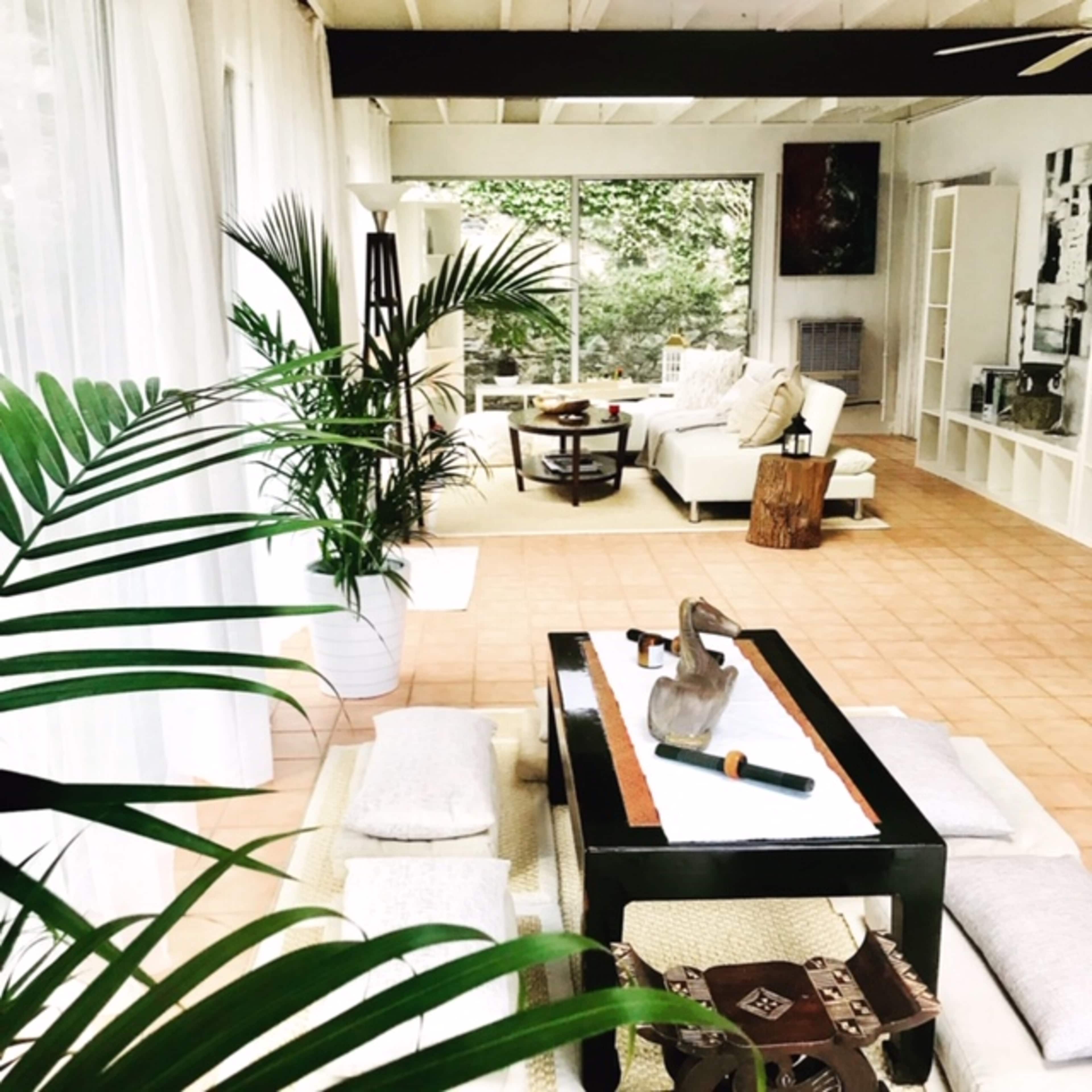 A spacious living room features white furniture, a low table, and large windows that open to a green outdoor space.