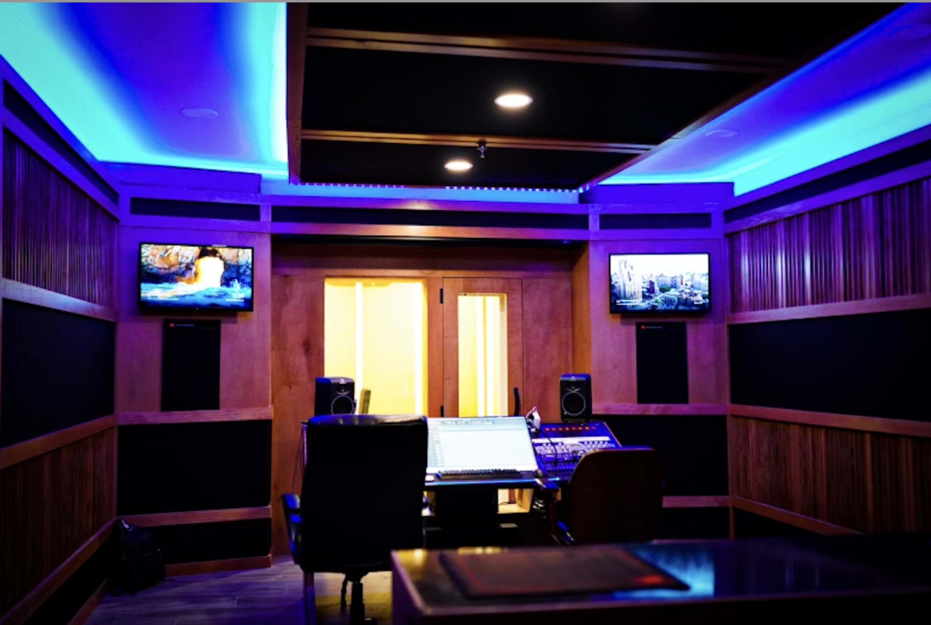 A music recording studio with two monitors, a mixing console, and blue LED lighting along the walls.
