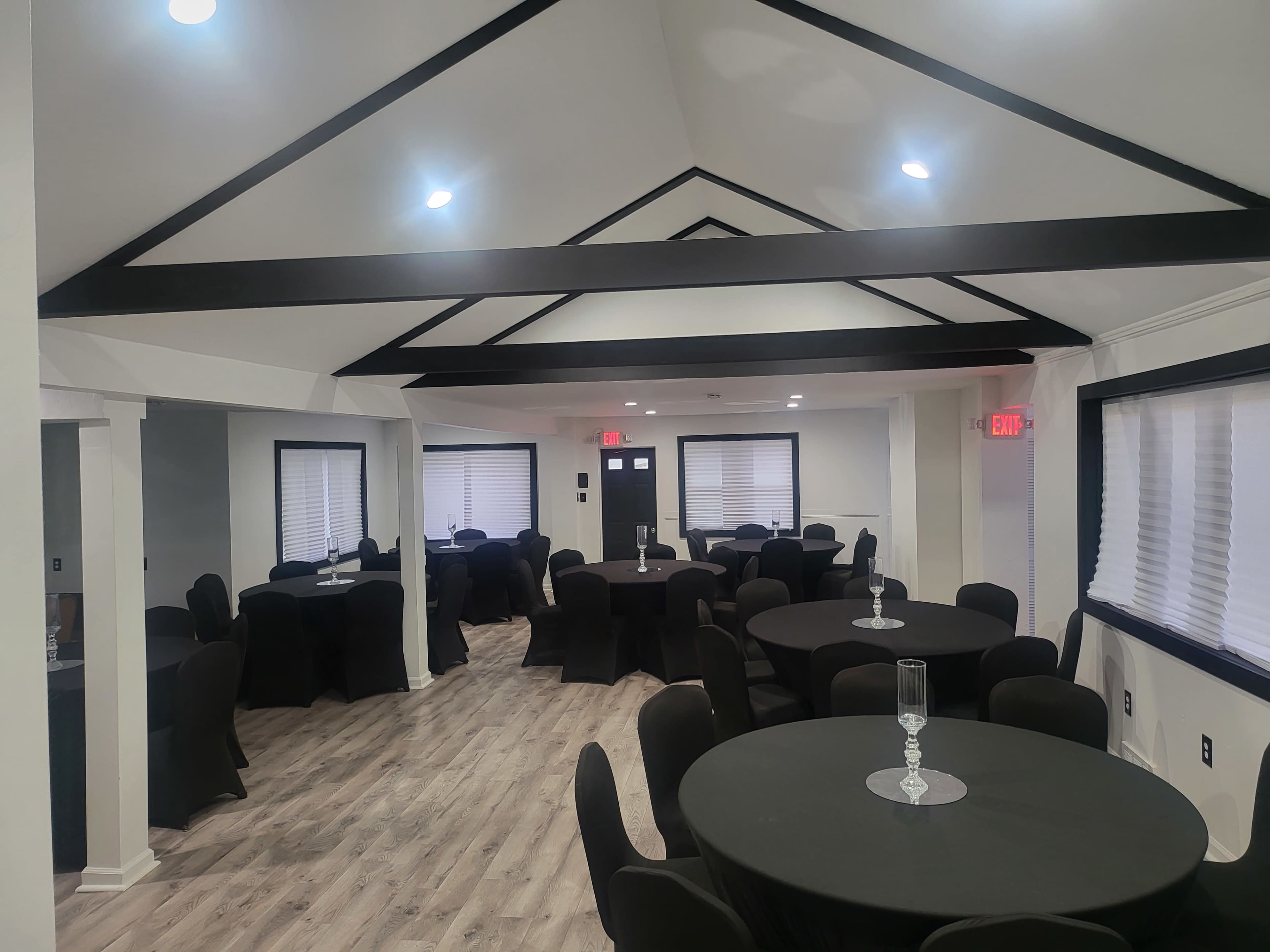 The image shows a spacious, well-lit event room featuring several round tables with black tablecloths and modern decor.