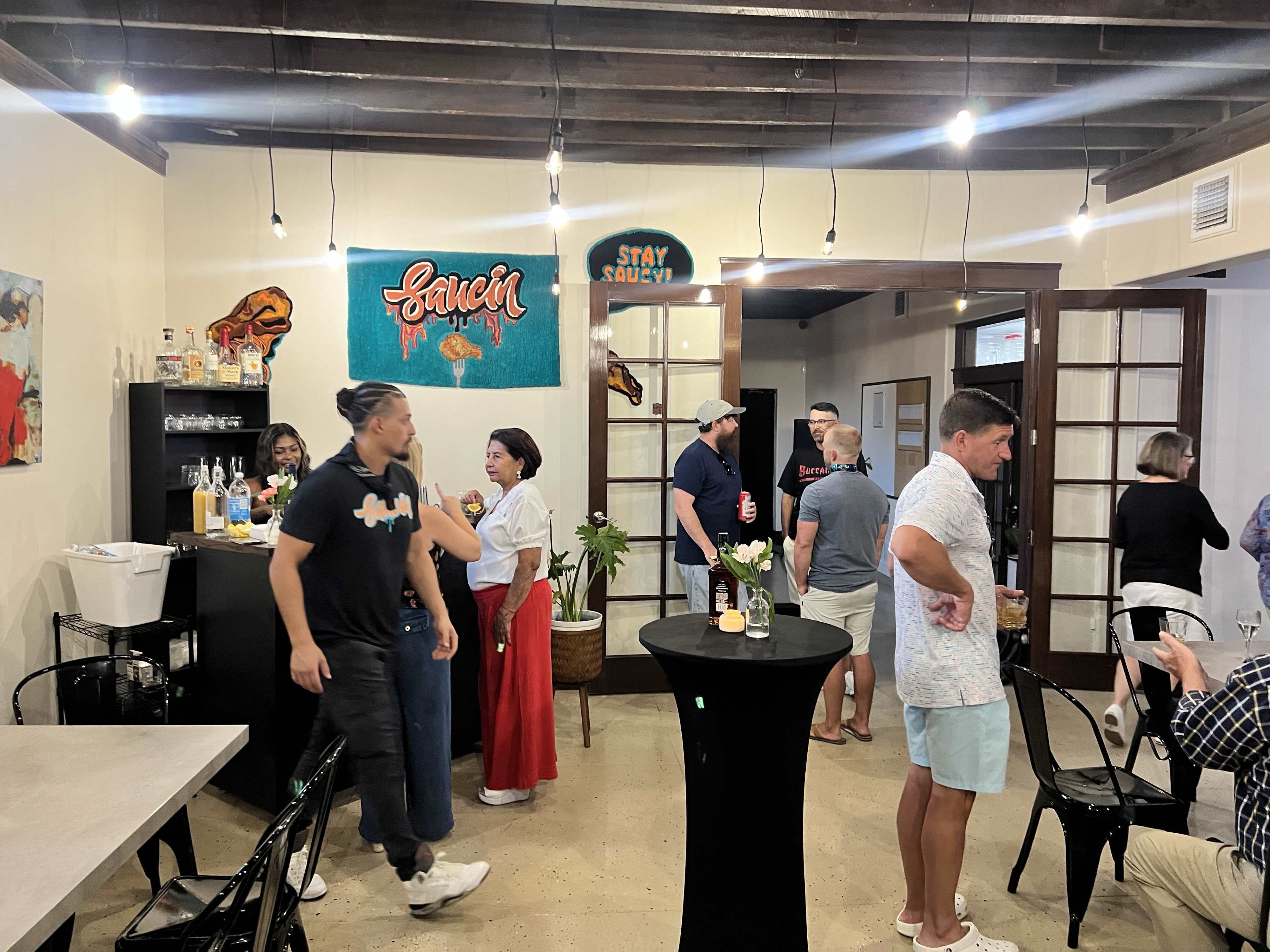 A group of people socializes in a well-lit event space featuring a bar, tables, and colorful wall decorations.