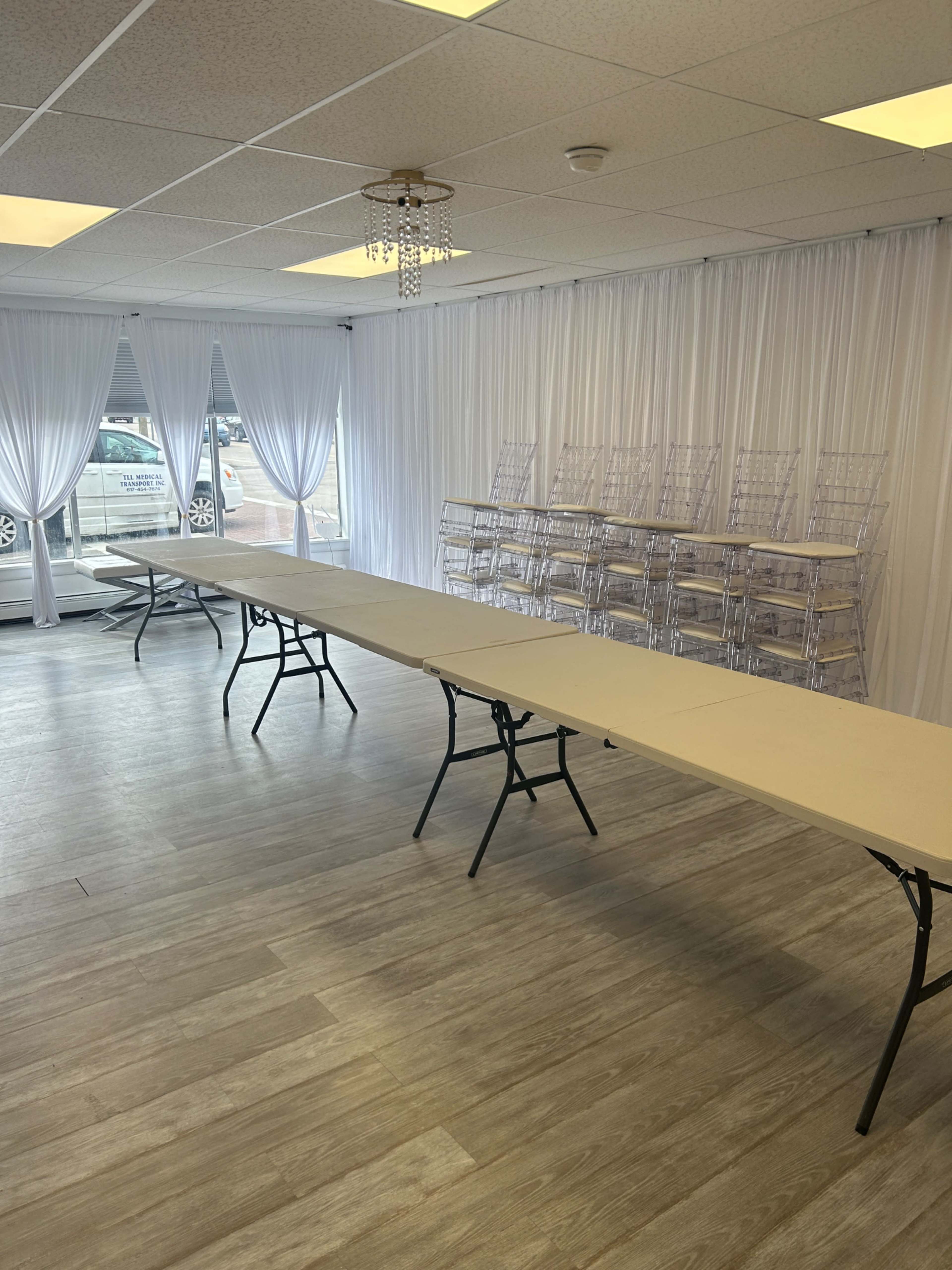 A spacious room features a long, narrow table and clear acrylic chairs stacked against a white curtain backdrop.