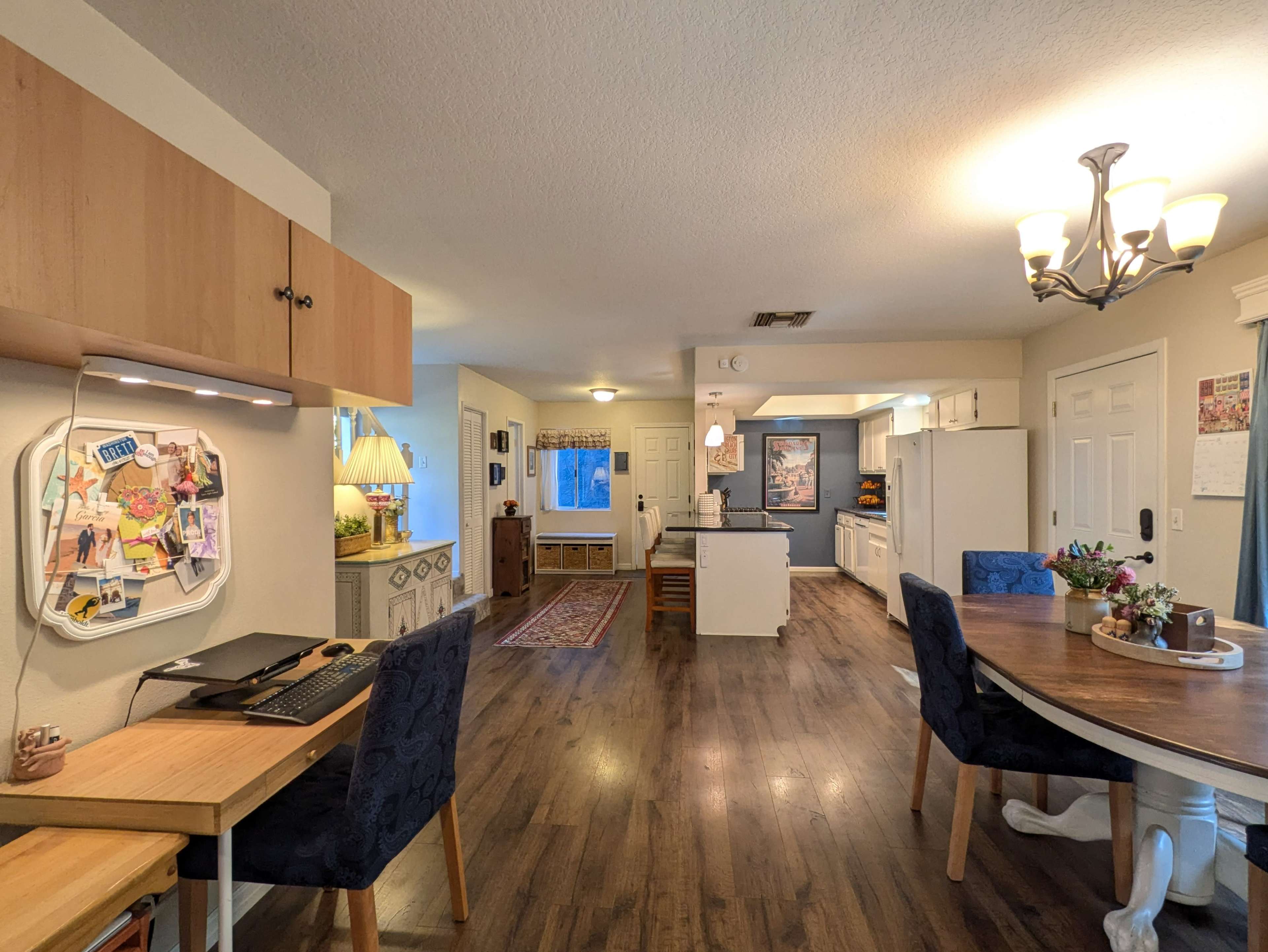 The image shows a spacious open-concept living area with a dining table, kitchen, and a workspace on one side, featuring hardwood floors and neutral-colored walls.