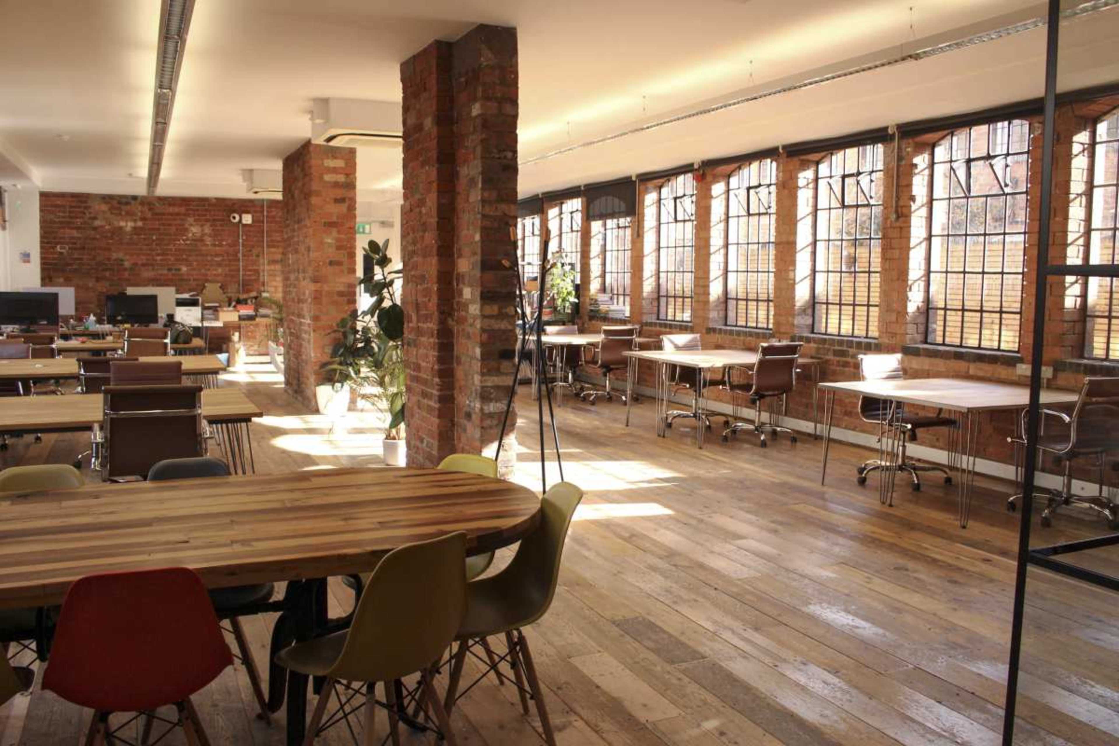 The image shows a spacious office environment with large windows, wooden floors, and various tables and chairs arranged throughout the room.