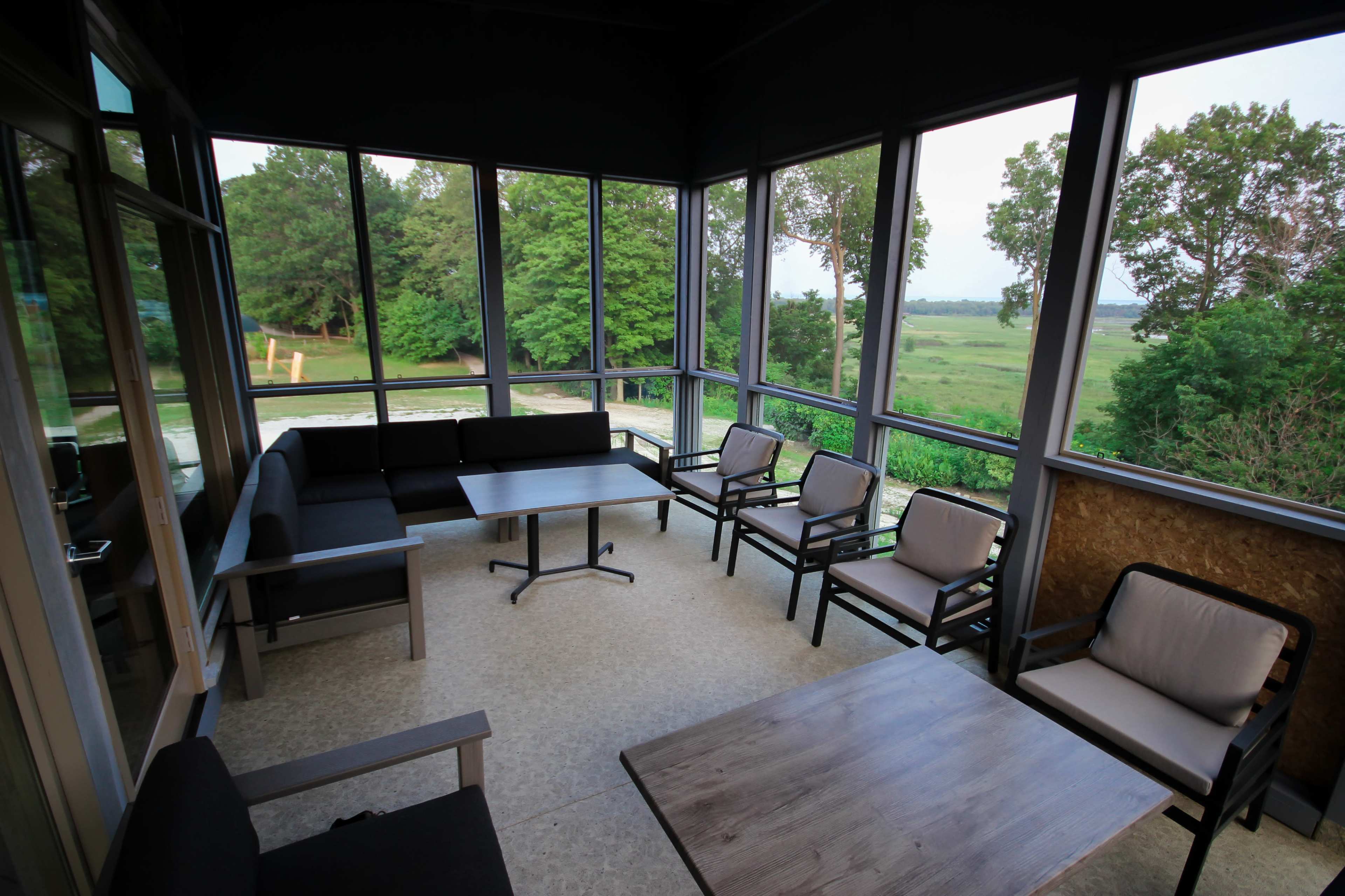 The image shows a screened-in porch with a seating area comprising a table and multiple chairs, overlooking a green landscape through large windows.