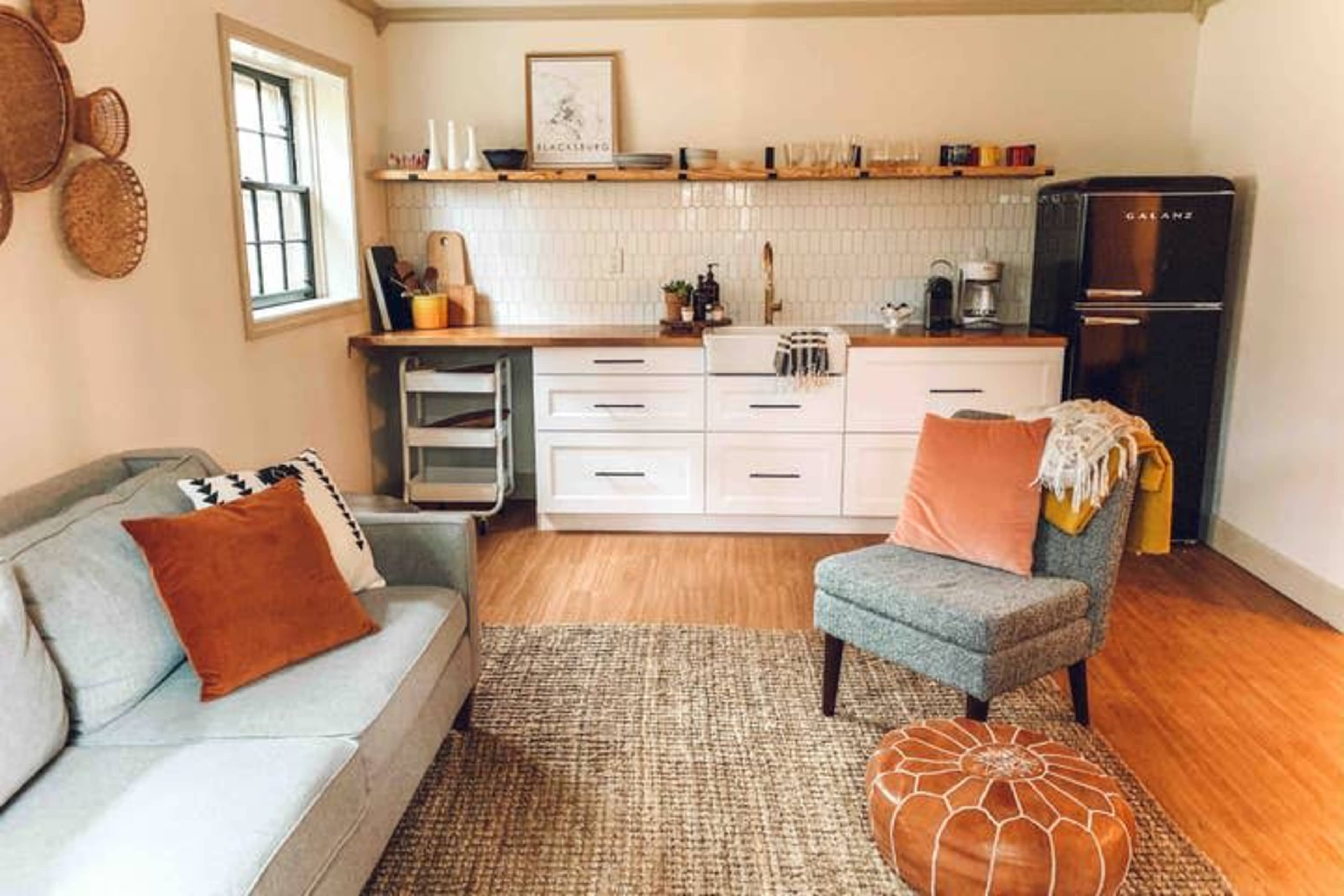 A modern kitchen and living area feature a white cabinetry, wooden countertop, and a gray couch with orange pillows, set on a woven rug.