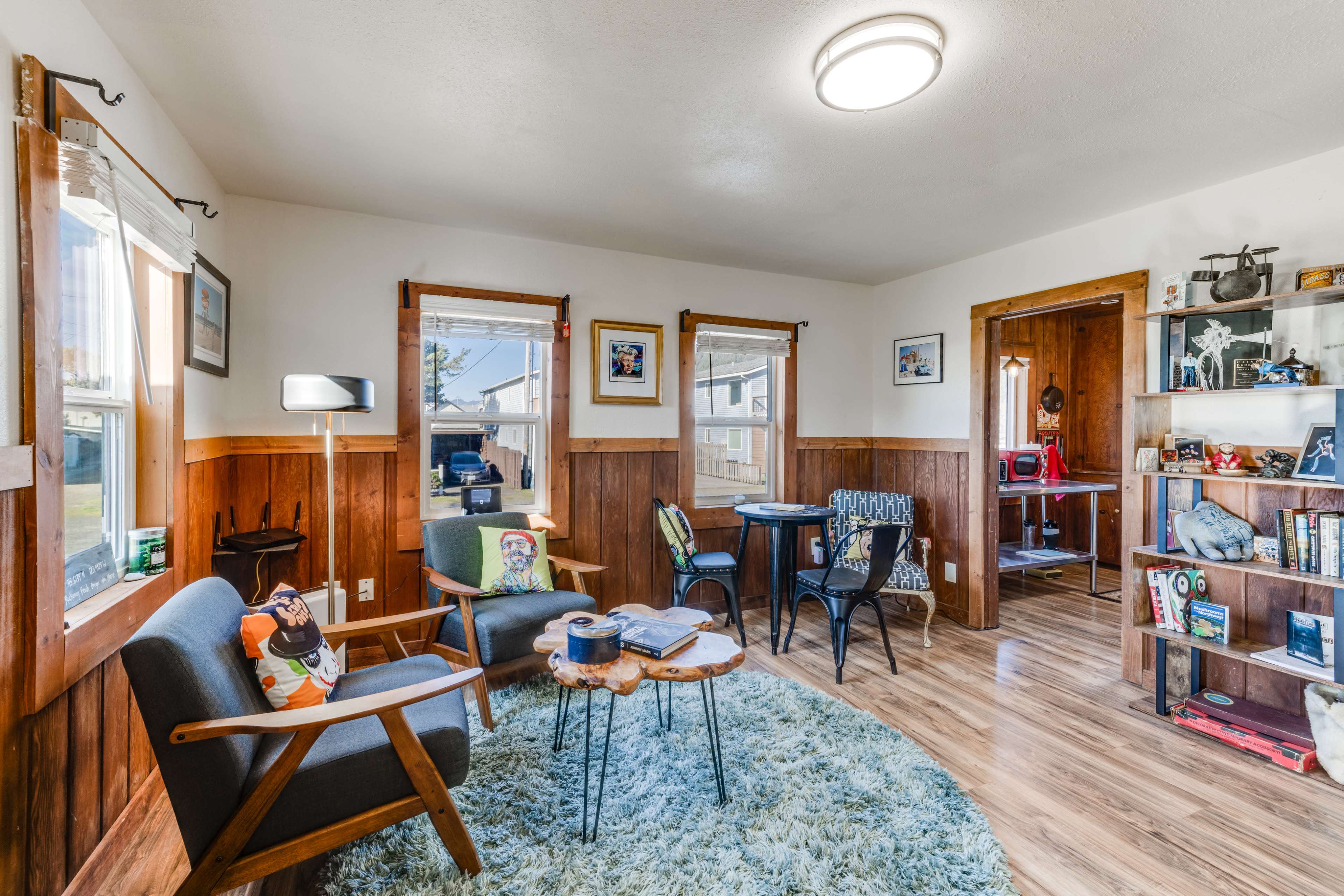 The image shows a cozy living room with wooden paneling, two chairs, a small table, and a bookshelf filled with various items.