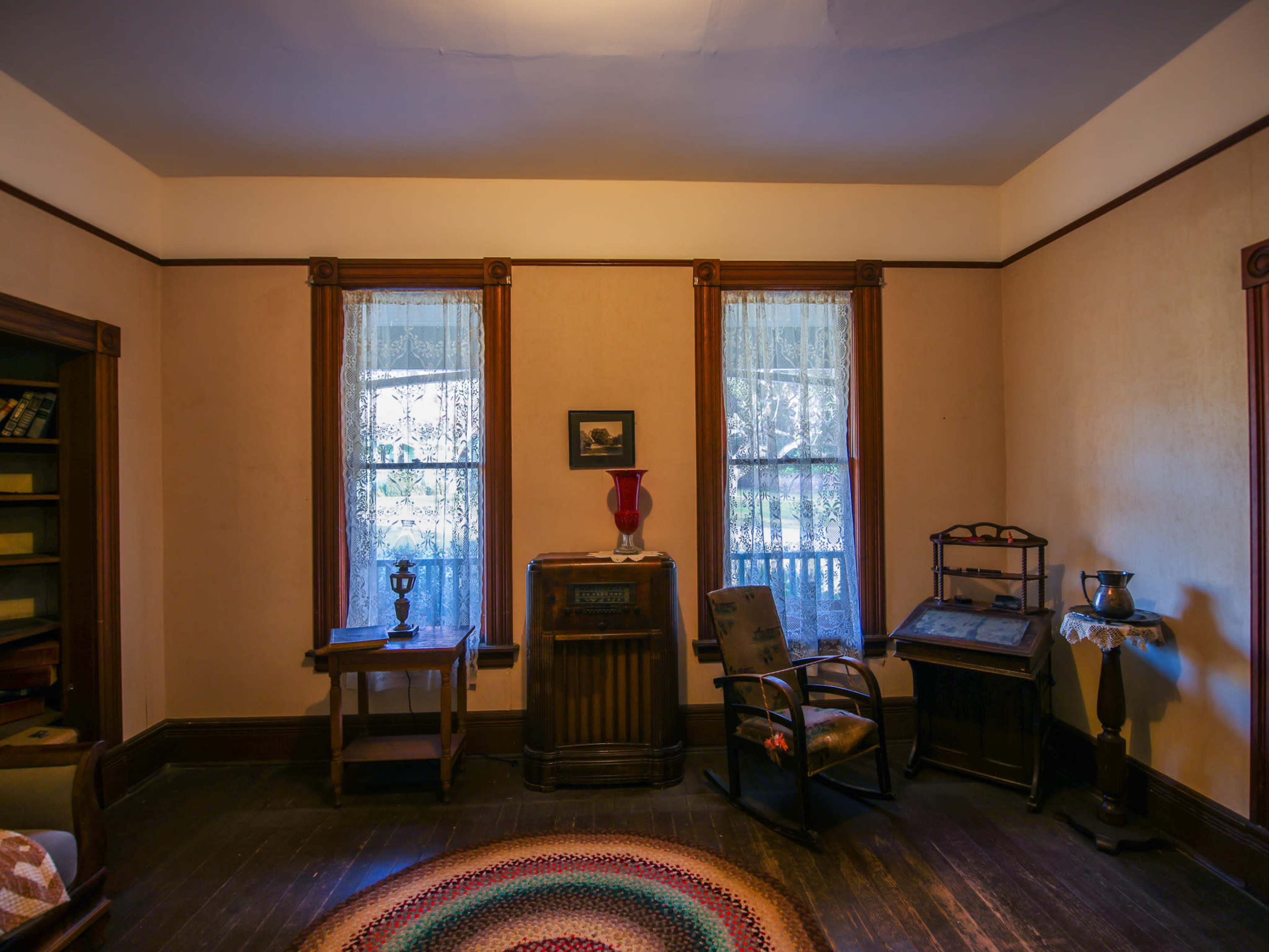 The room features two windows with lace curtains, a small table, a vintage radio, and a rocking chair, all arranged around a circular rug.