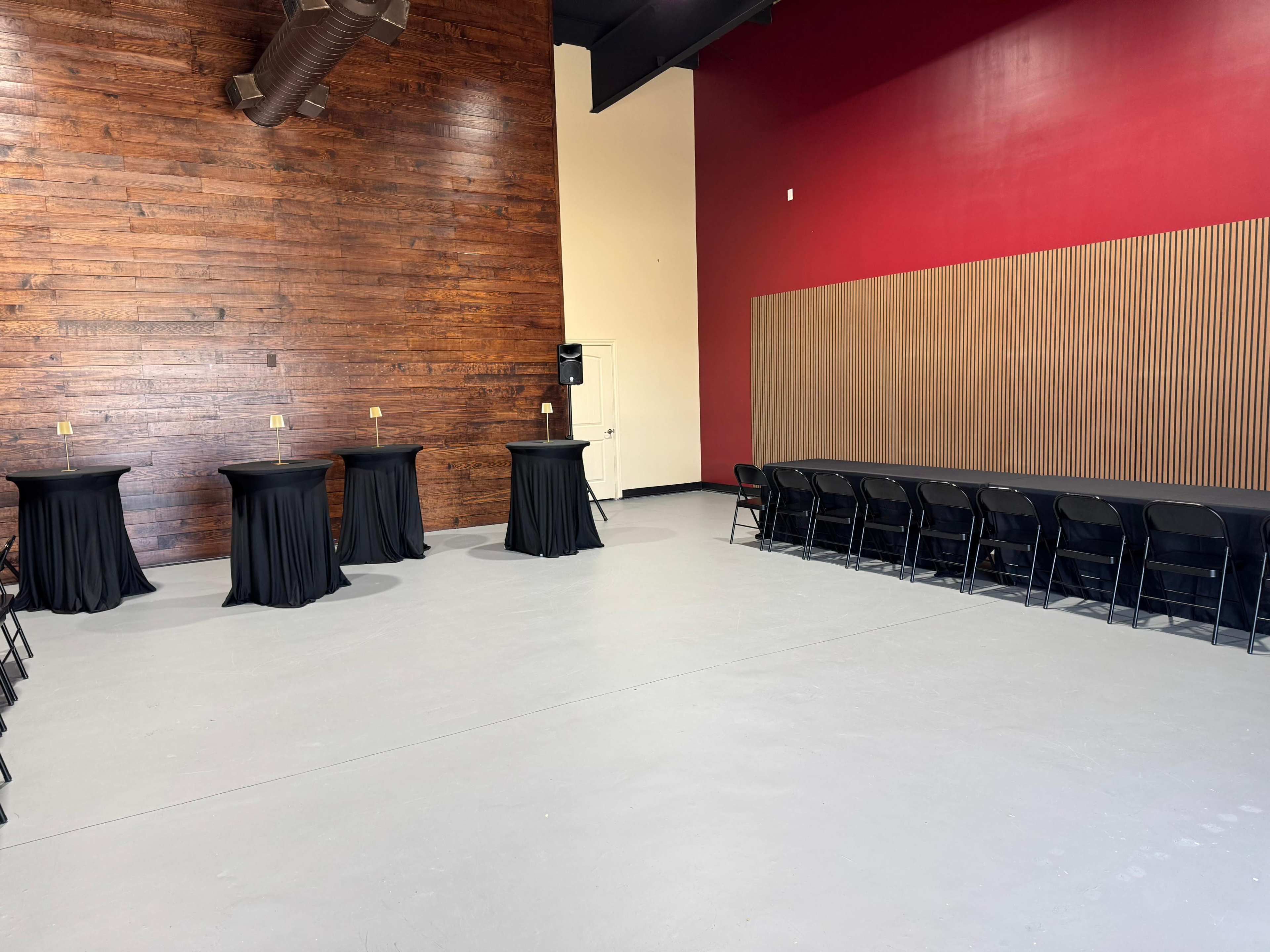 The image shows an empty event space featuring a wooden wall, a red accent wall, black tables with black tablecloths, and a row of black chairs against one side.