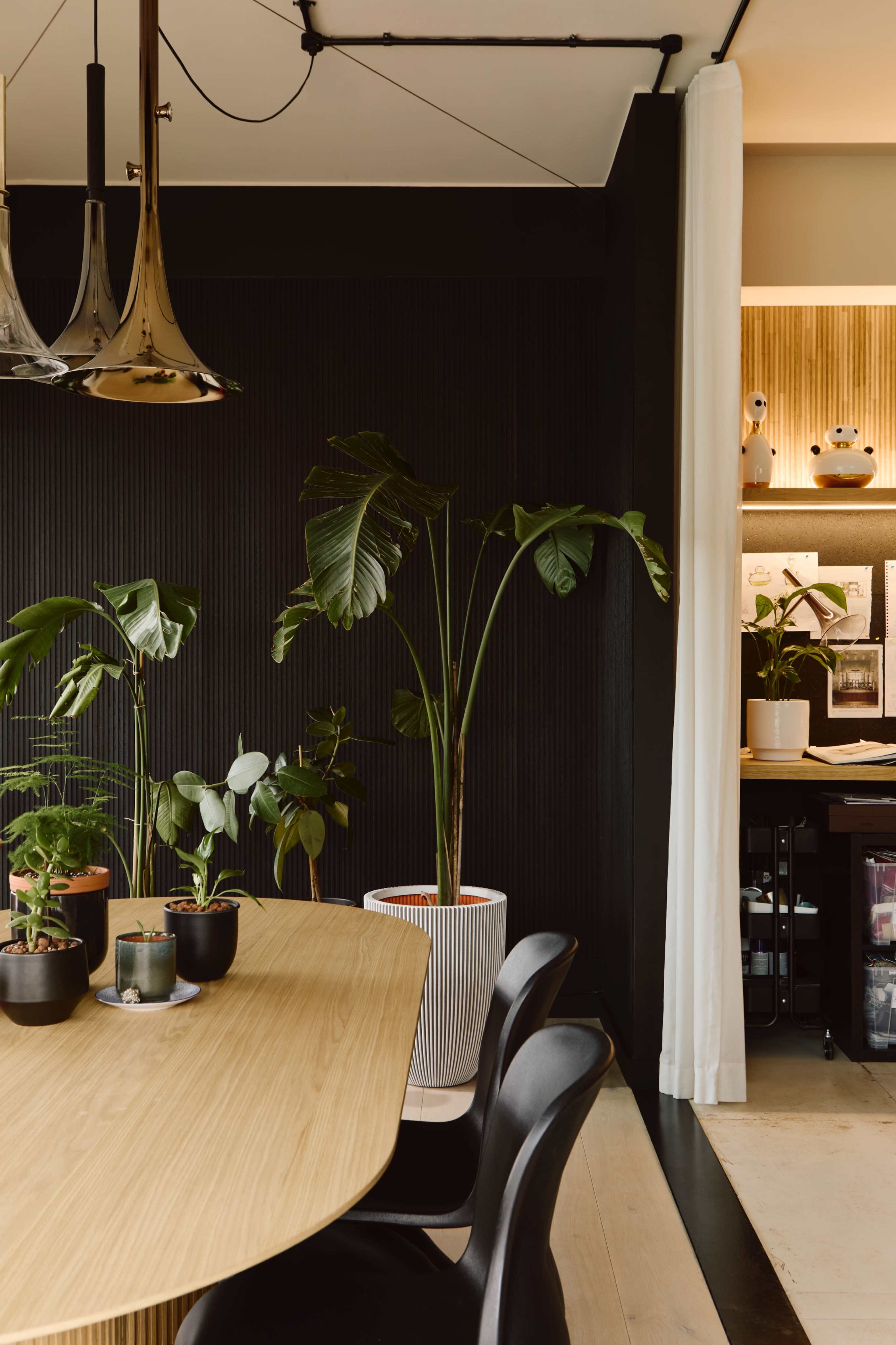 A modern dining area features a round wooden table surrounded by black chairs, with potted plants on the table and in a large planter against a textured black wall.