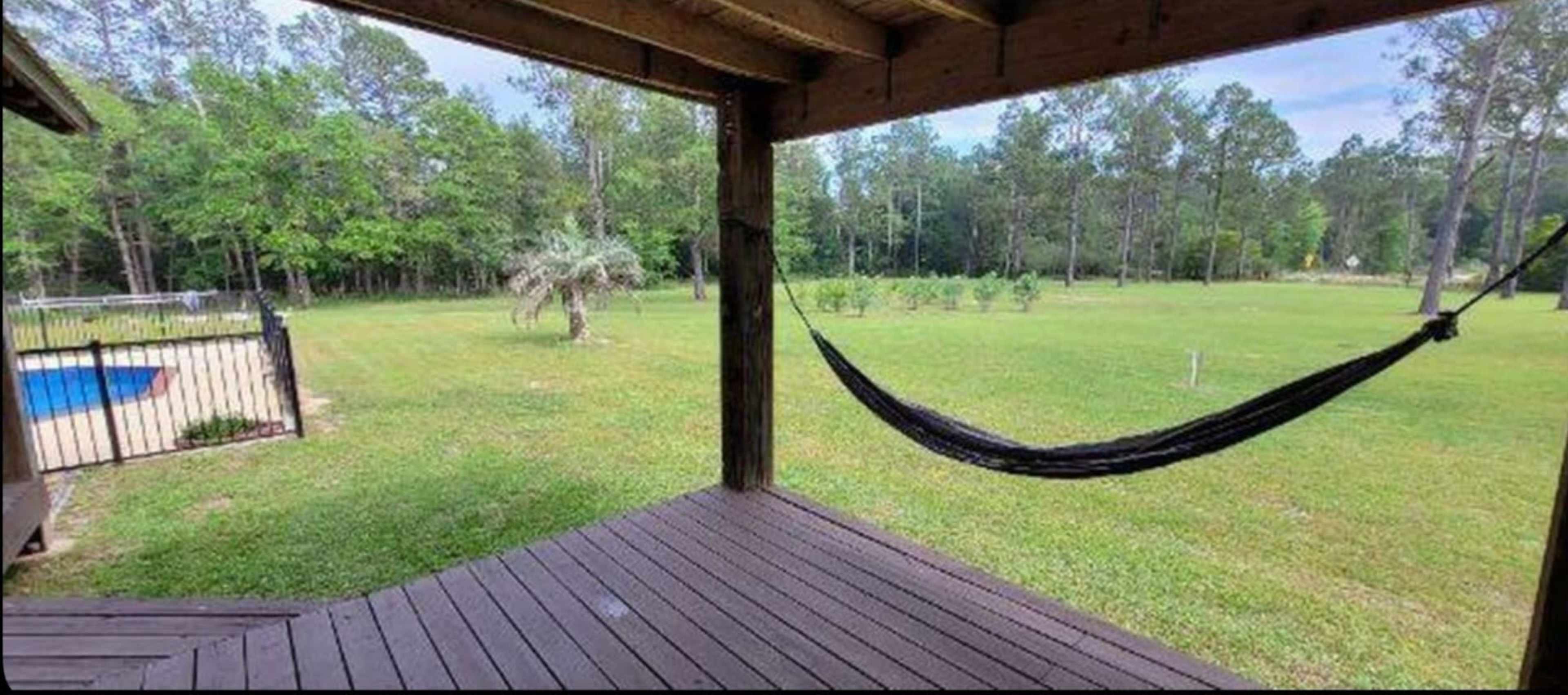 A hammock is suspended from a porch overlooking a grassy yard with trees and a pool in the background.
