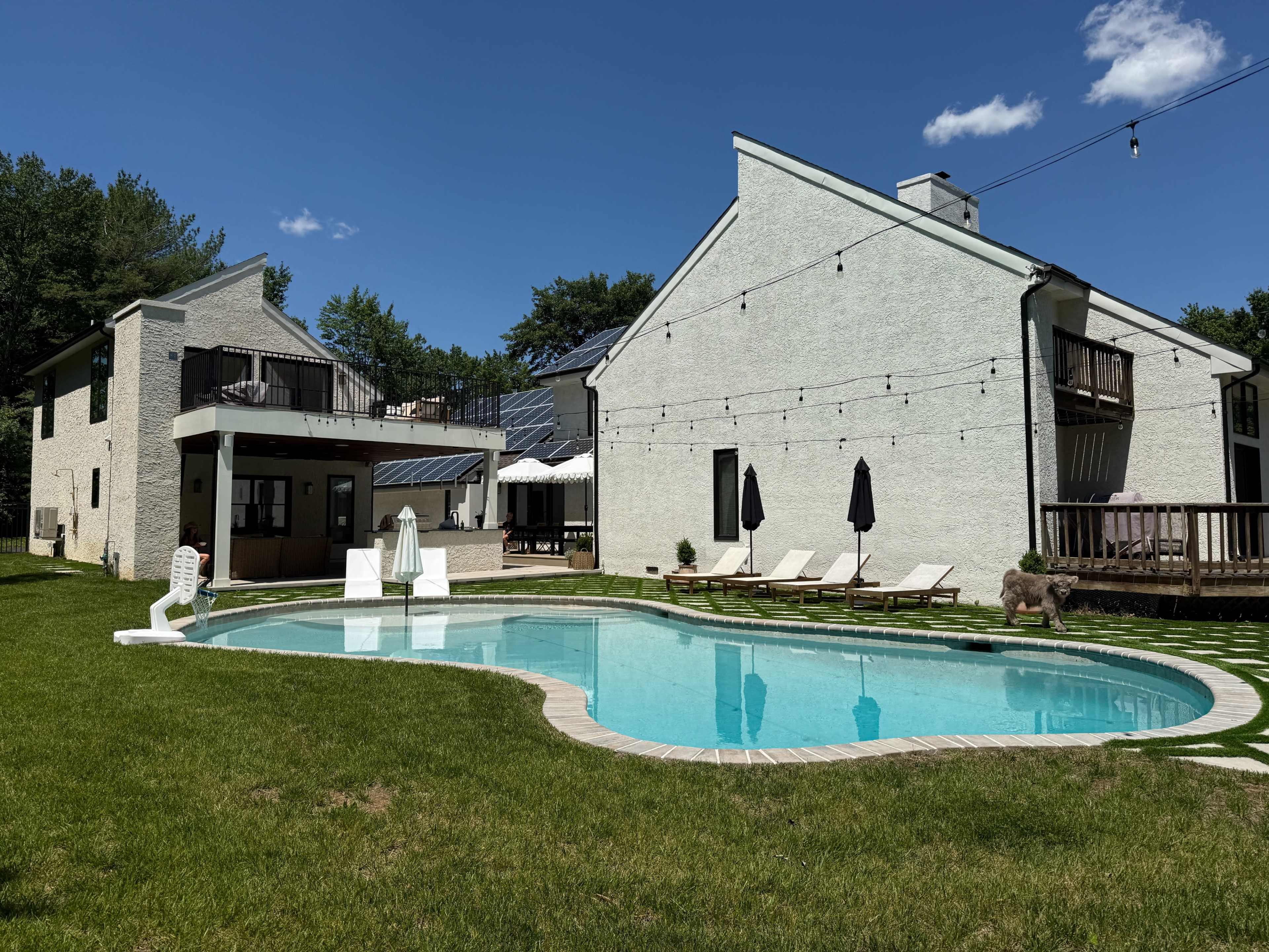 The image shows a residential property with a swimming pool in the foreground, surrounded by grass, and two white houses in the background under a clear blue sky.