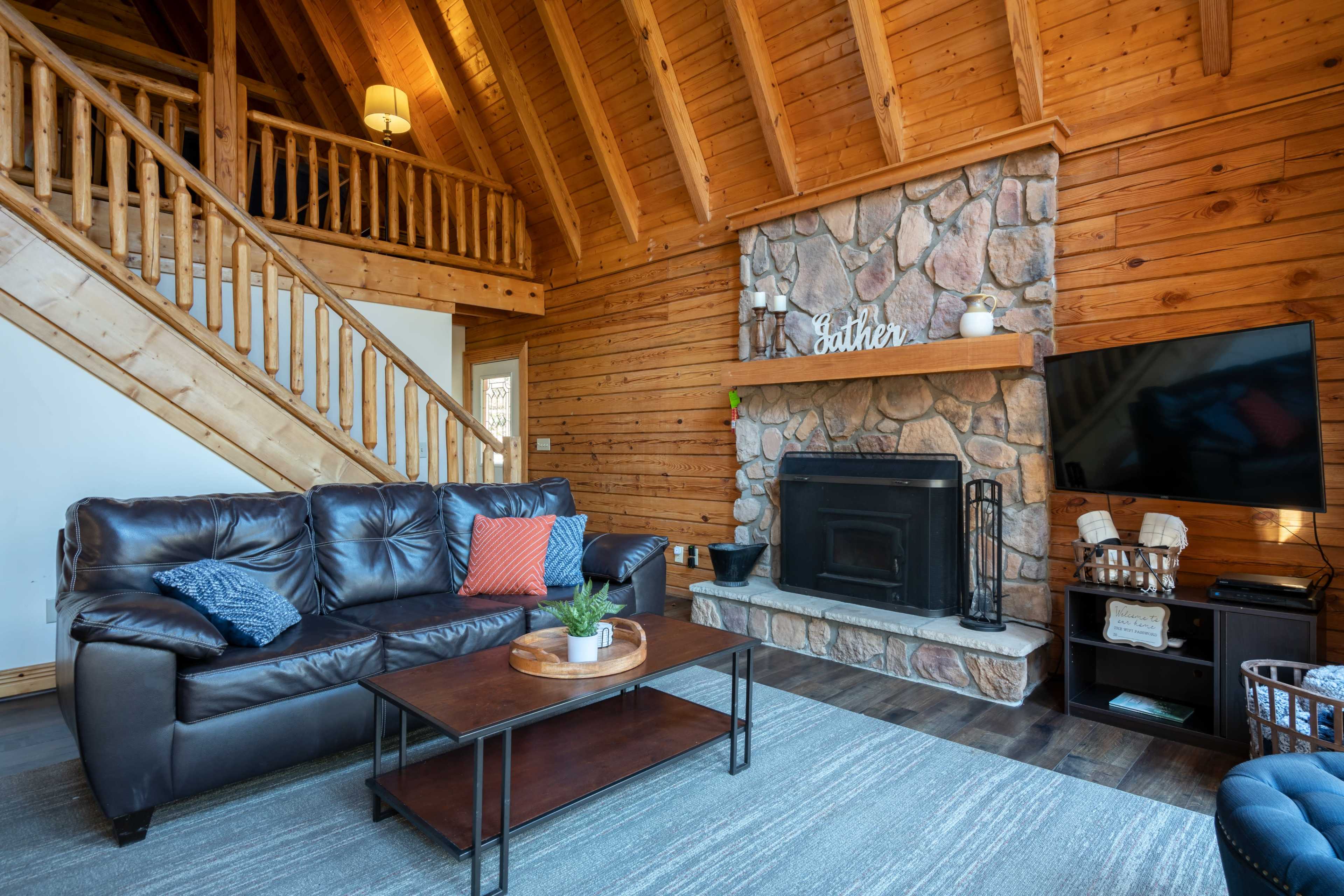 A cozy living room featuring a stone fireplace, wooden beams, and a leather sofa arranged with decorative cushions.