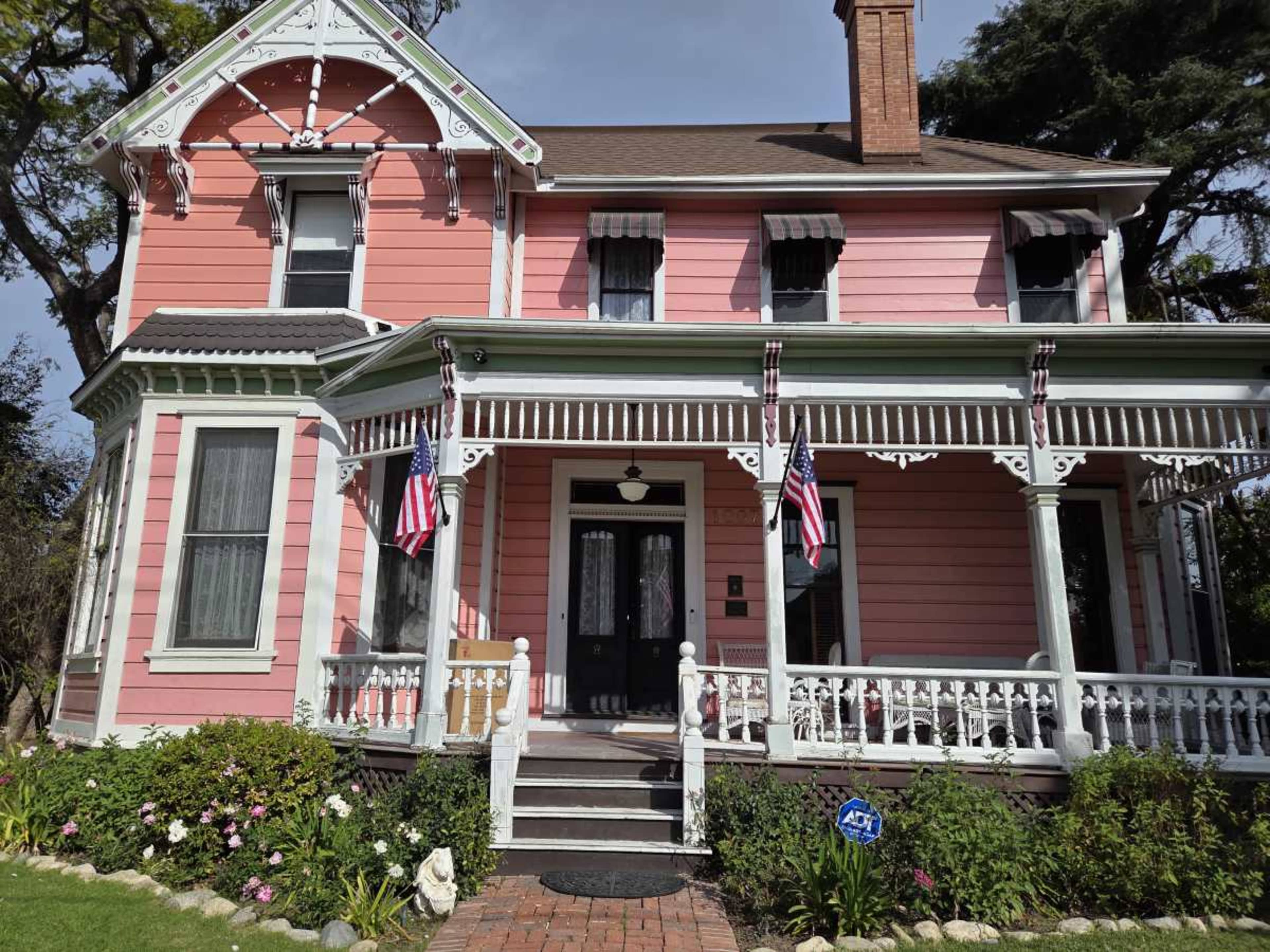 A pink Victorian house features intricate decorative elements, a wide front porch with white railings, and two American flags hanging from the porch.