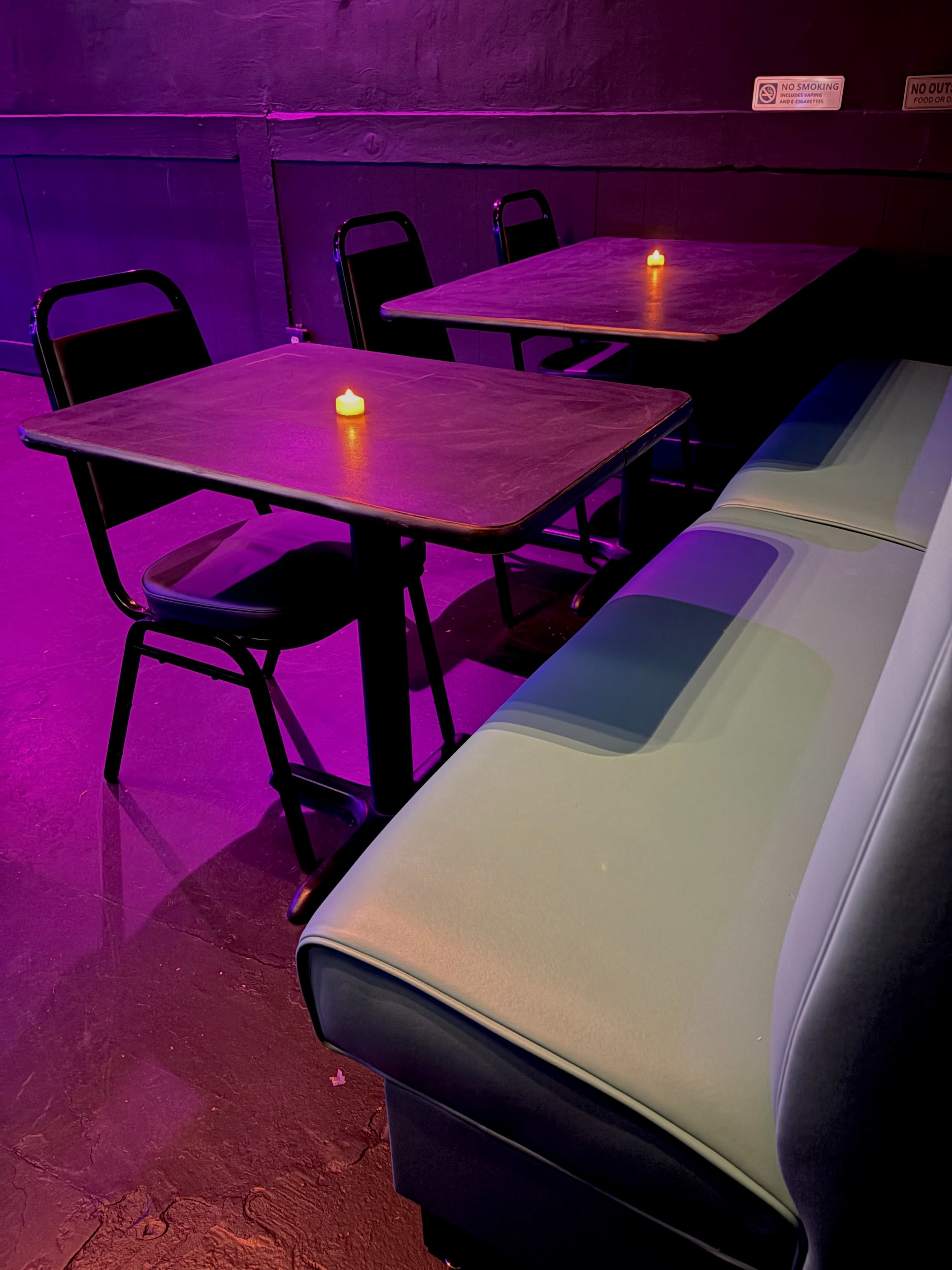 The image shows a dimly lit seating area with two tables, one made of dark wood, surrounded by black chairs and a cushioned green bench, illuminated by the soft glow of a candle on each table.