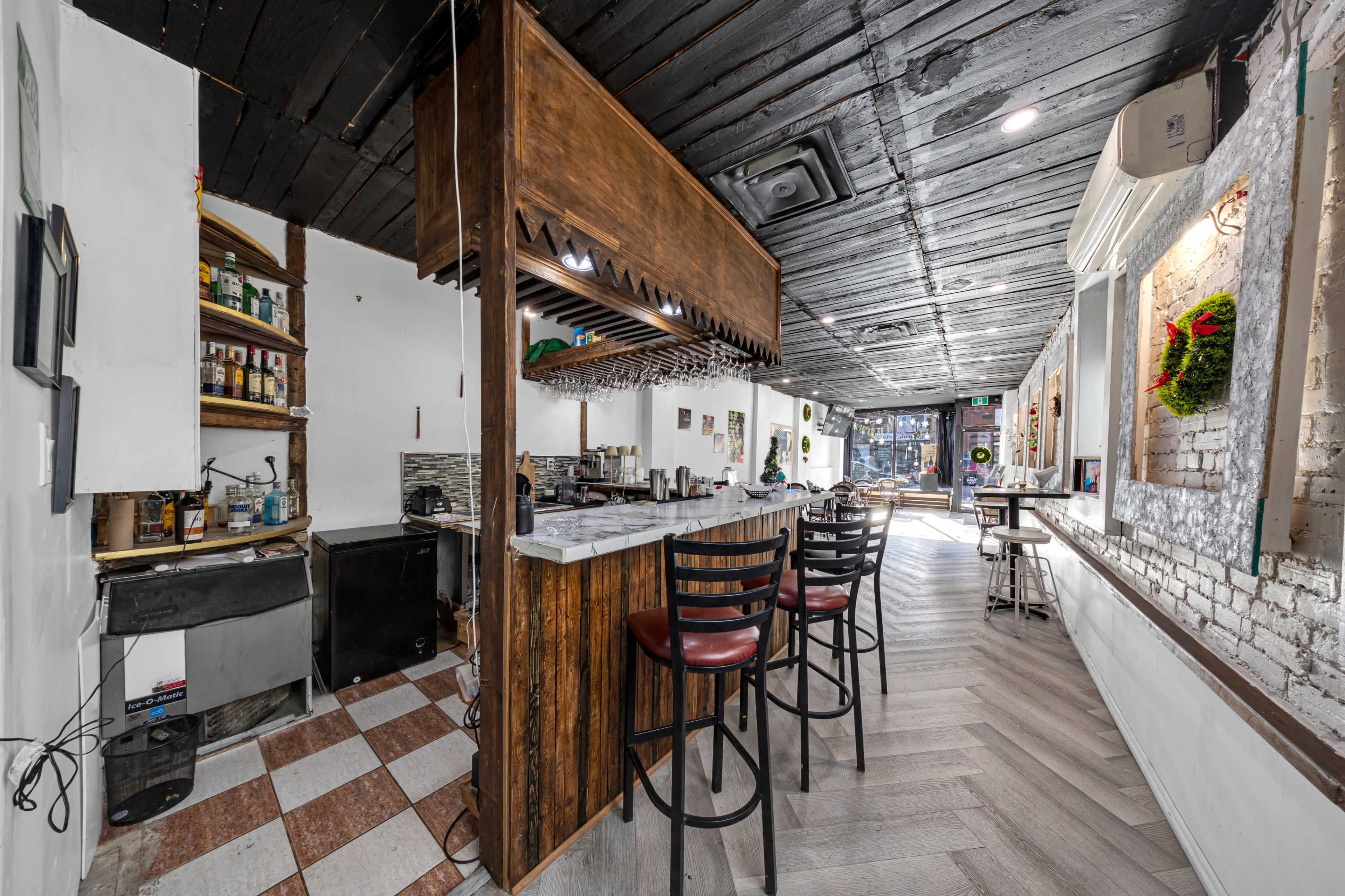 The image shows a dimly lit cafe or bar interior featuring a wooden counter with high stools, shelves stocked with bottles, and a tiled floor.