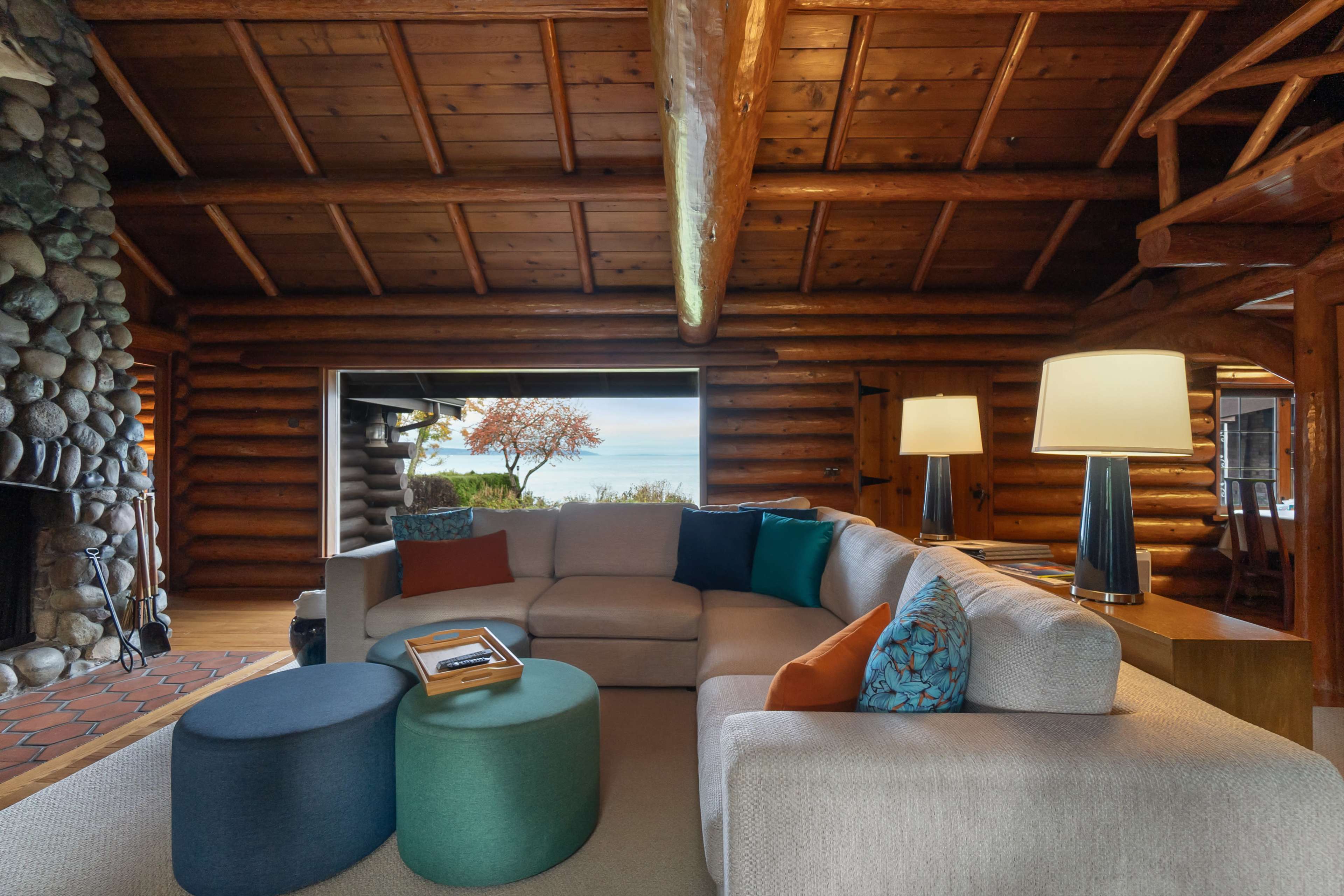 The image shows a spacious log cabin interior featuring a sectional sofa with colorful cushions, a stone fireplace, and large windows that open to a scenic view.
