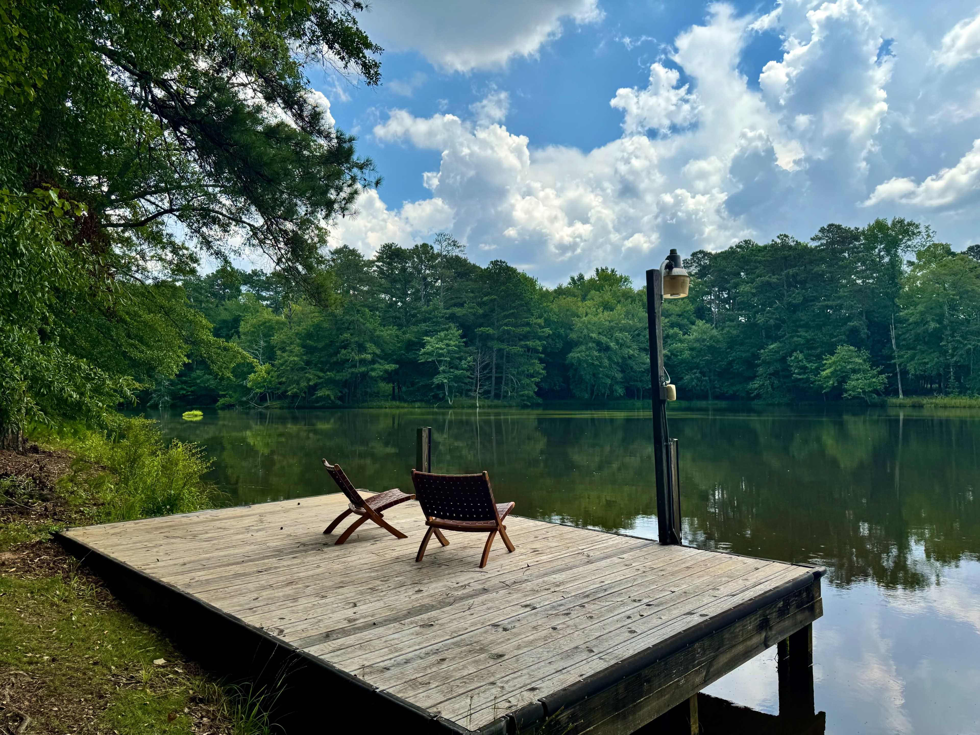 Two wooden chairs sit on a fishing dock overlooking a calm lake surrounded by trees under a partly cloudy sky.