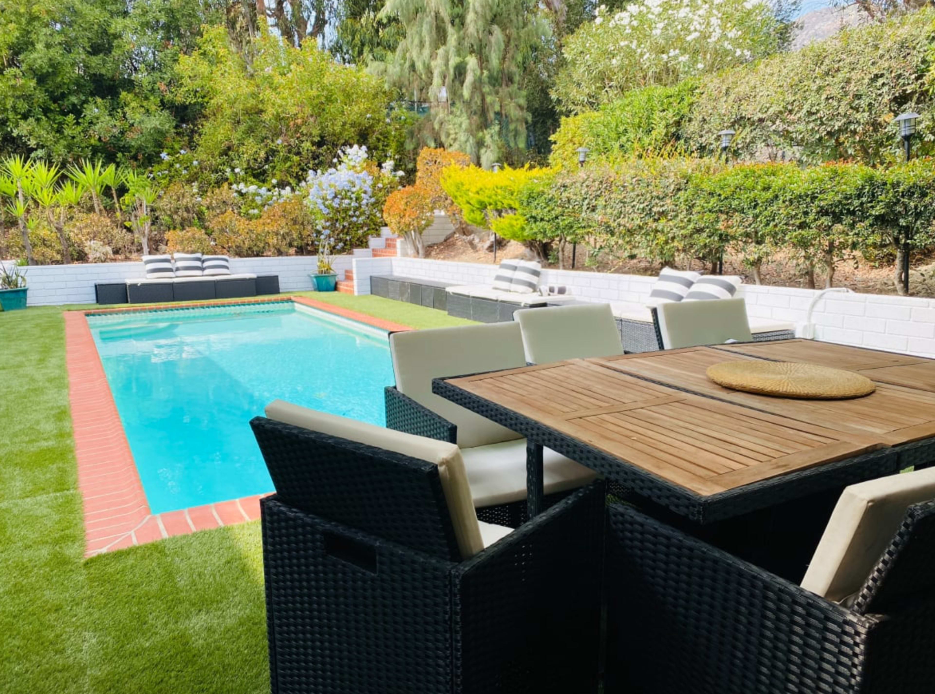 A backyard with a swimming pool, surrounded by greenery, and a dining table set with chairs nearby.