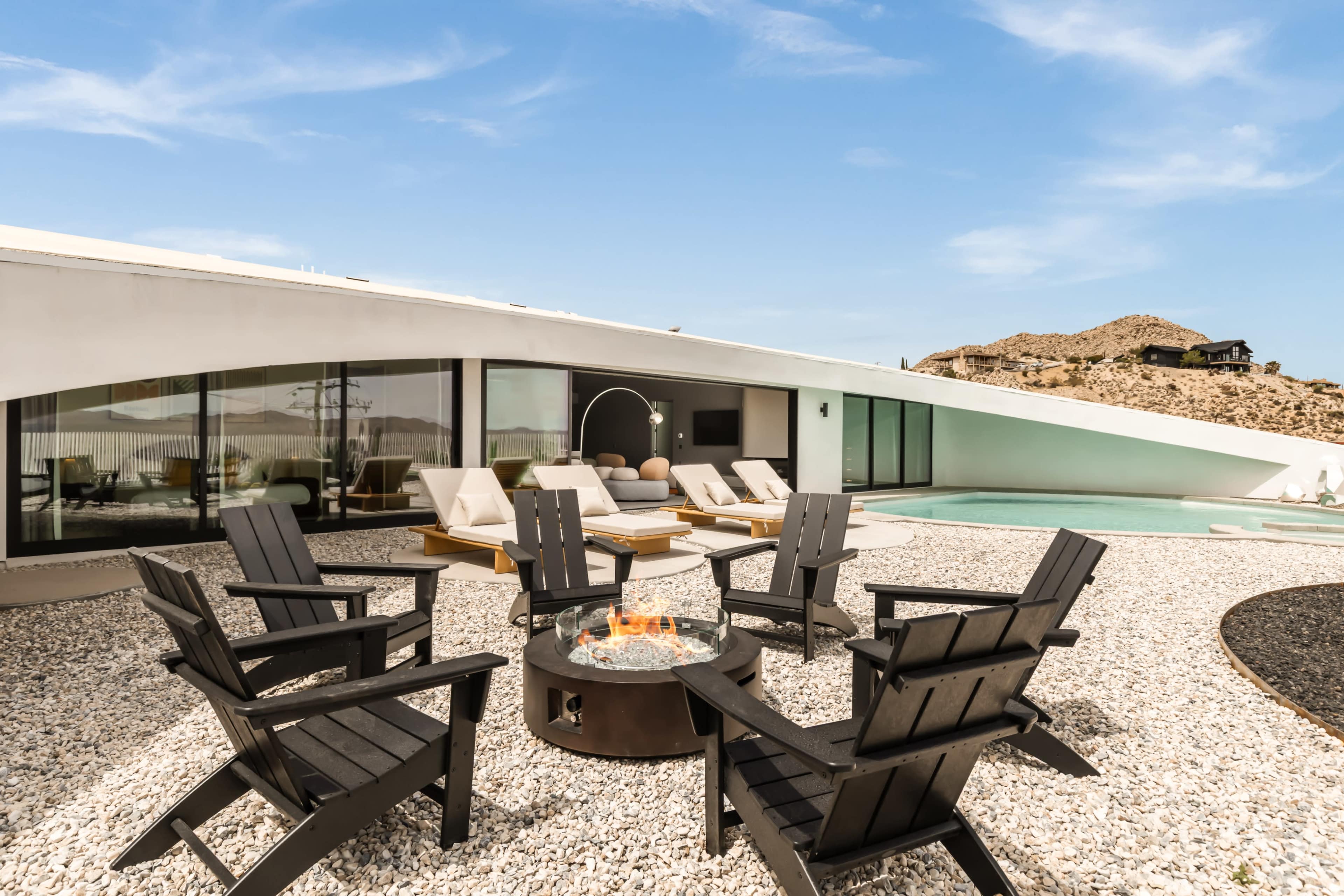 A modern outdoor seating area features black Adirondack chairs arranged around a fire pit, with a sleek white building and a pool in the background against a mountainous landscape.