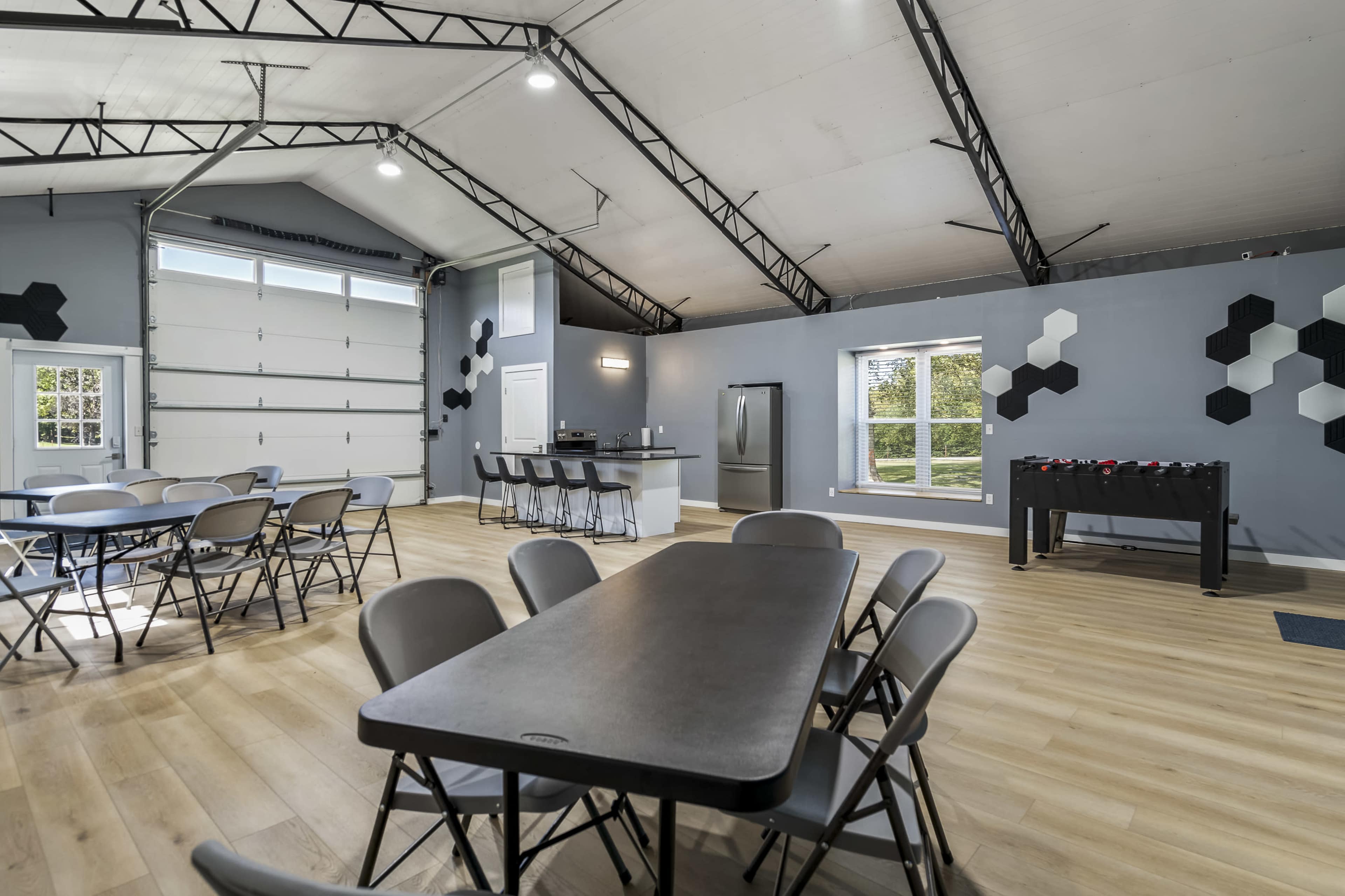 The image shows an interior space with high ceilings, featuring metal folding tables and chairs, a kitchenette area, and a foosball table against a wall.