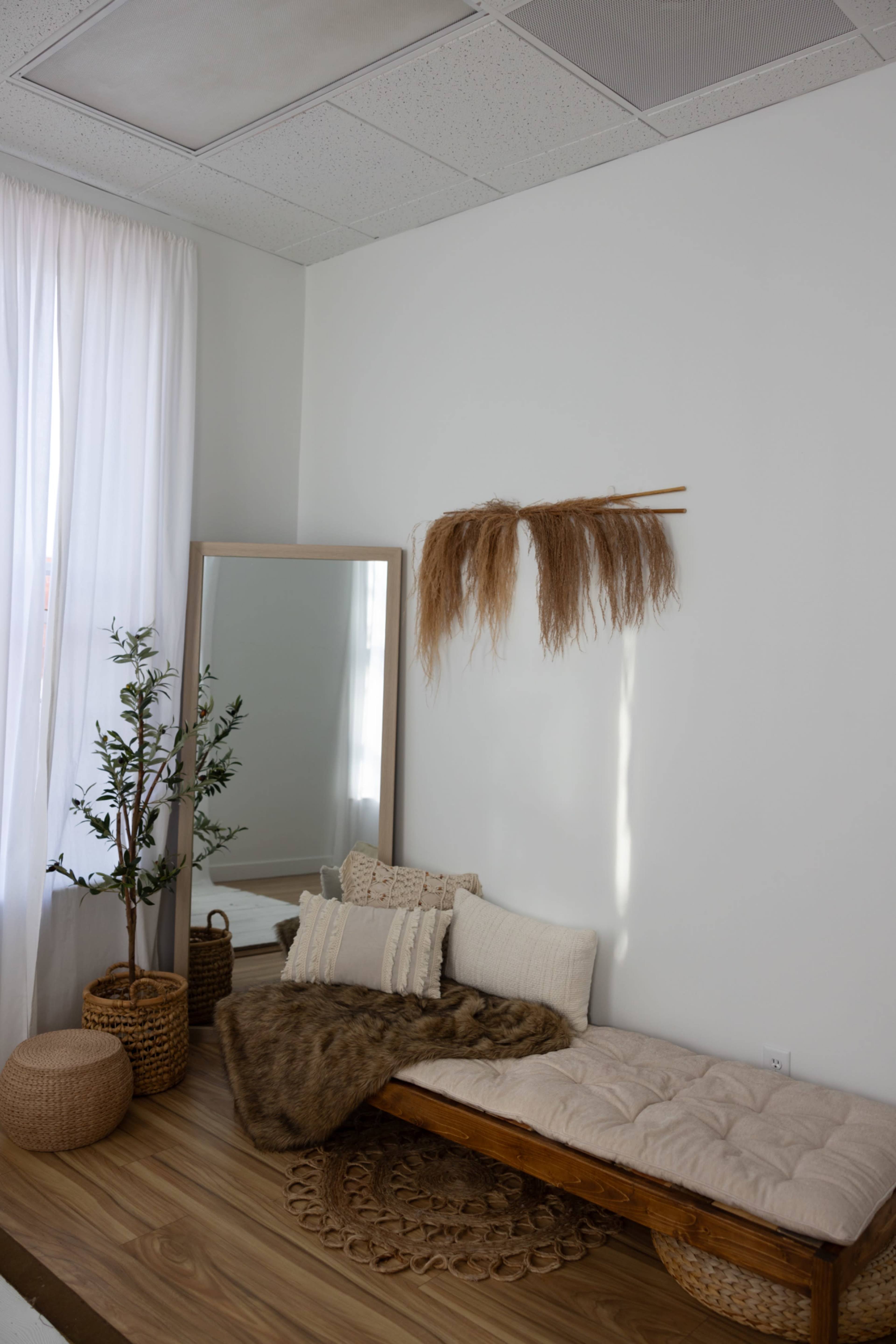 A minimalist interior features a wooden bench with cushions, a large mirror, a potted plant, and decorative items on a wooden floor.