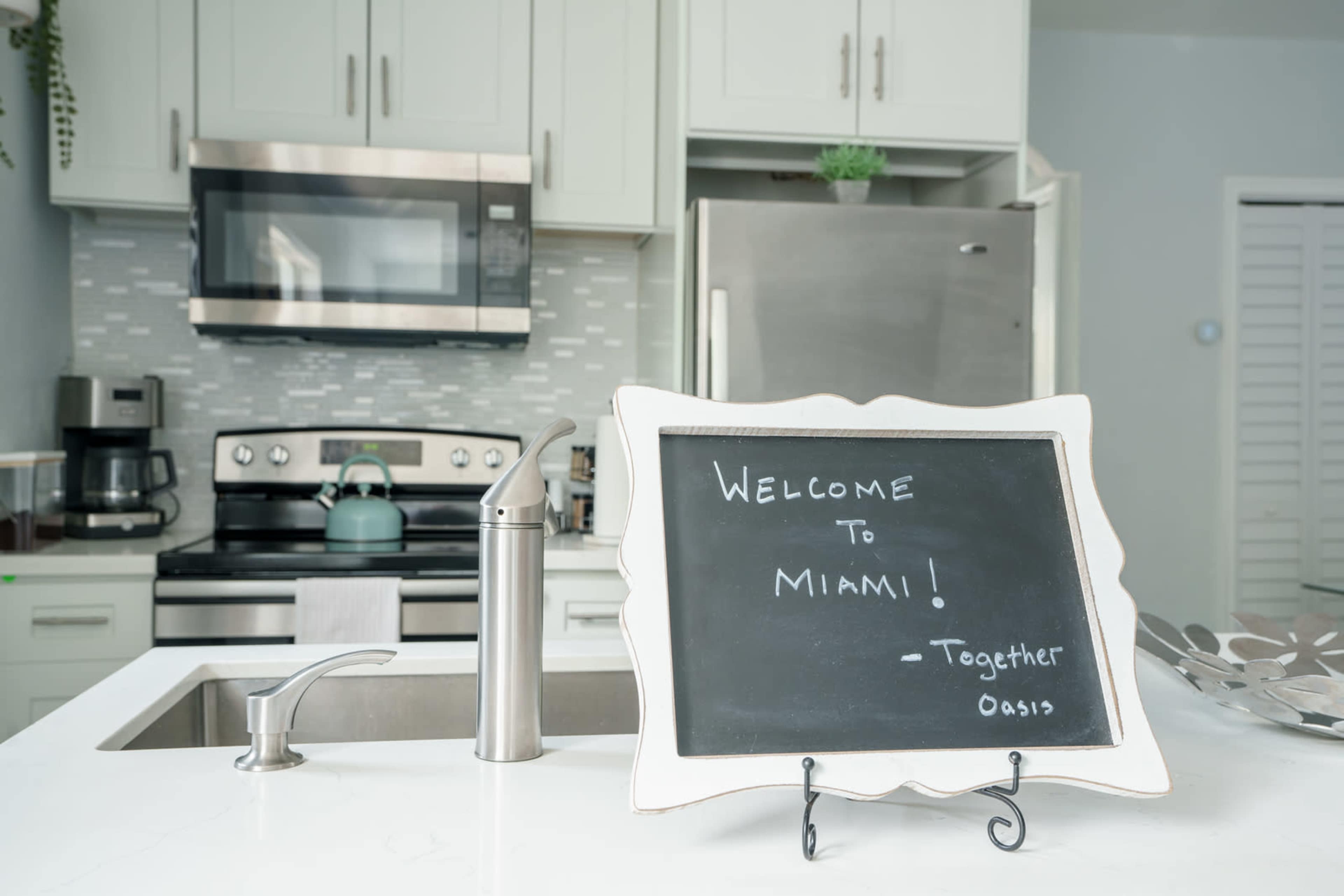 A chalkboard sign reading "WELCOME TO MIAMI!" stands in the foreground of a modern kitchen featuring stainless steel appliances and a sleek countertop.