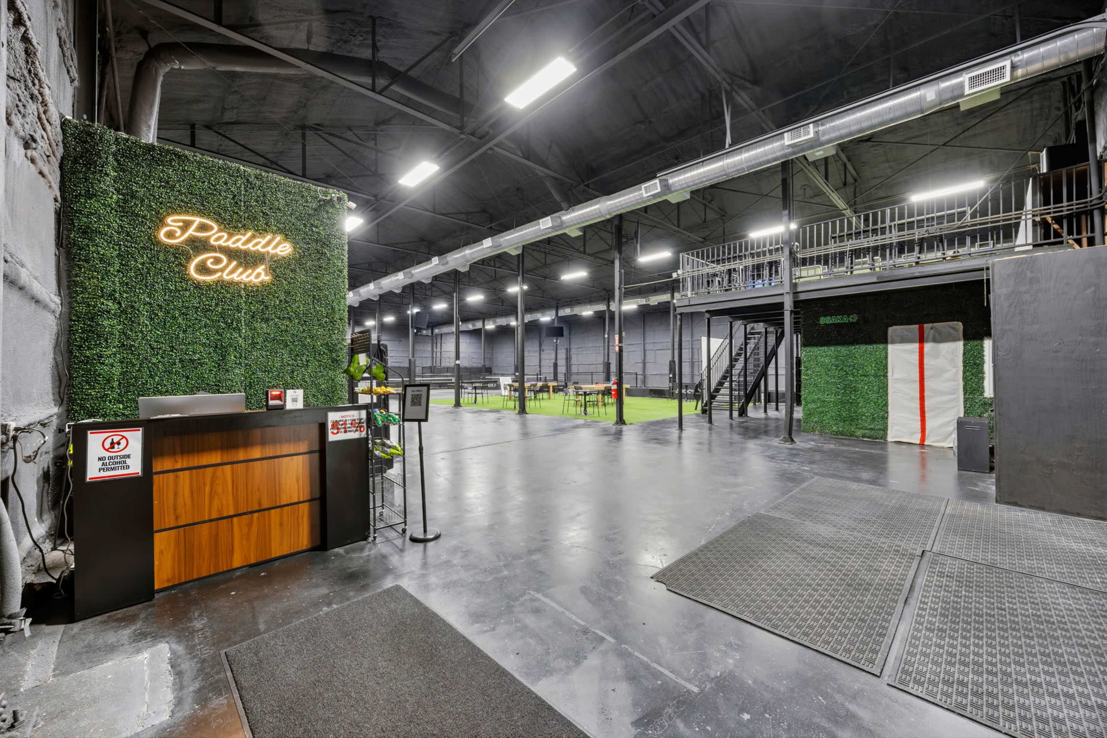The image depicts an indoor sports facility with a reception area featuring greenery and signage, alongside a spacious playing field and elevated viewing area.
