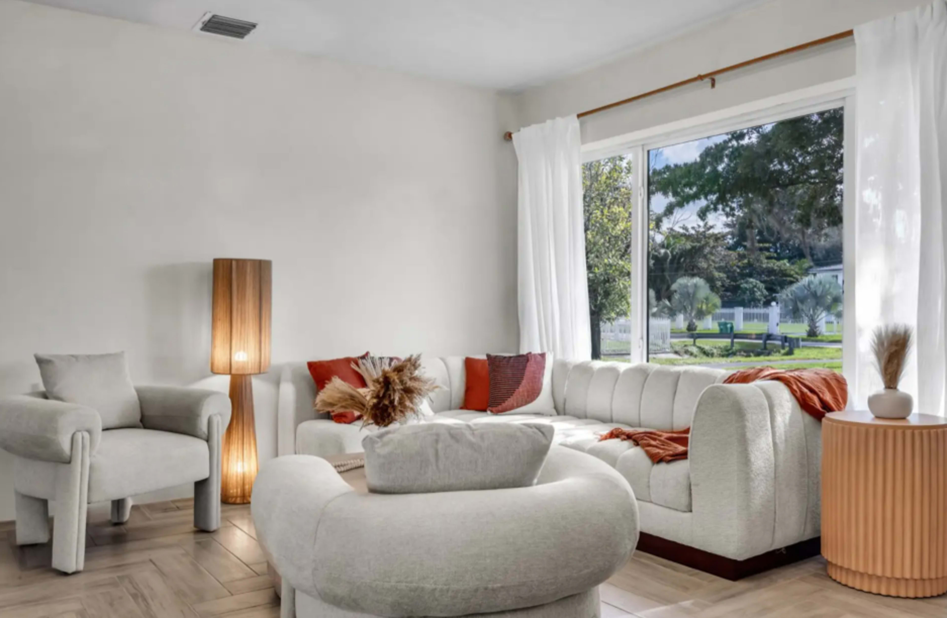 The image shows a modern living room with a light-colored sectional sofa, two armchairs, and a round side table, illuminated by natural light from a large window.
