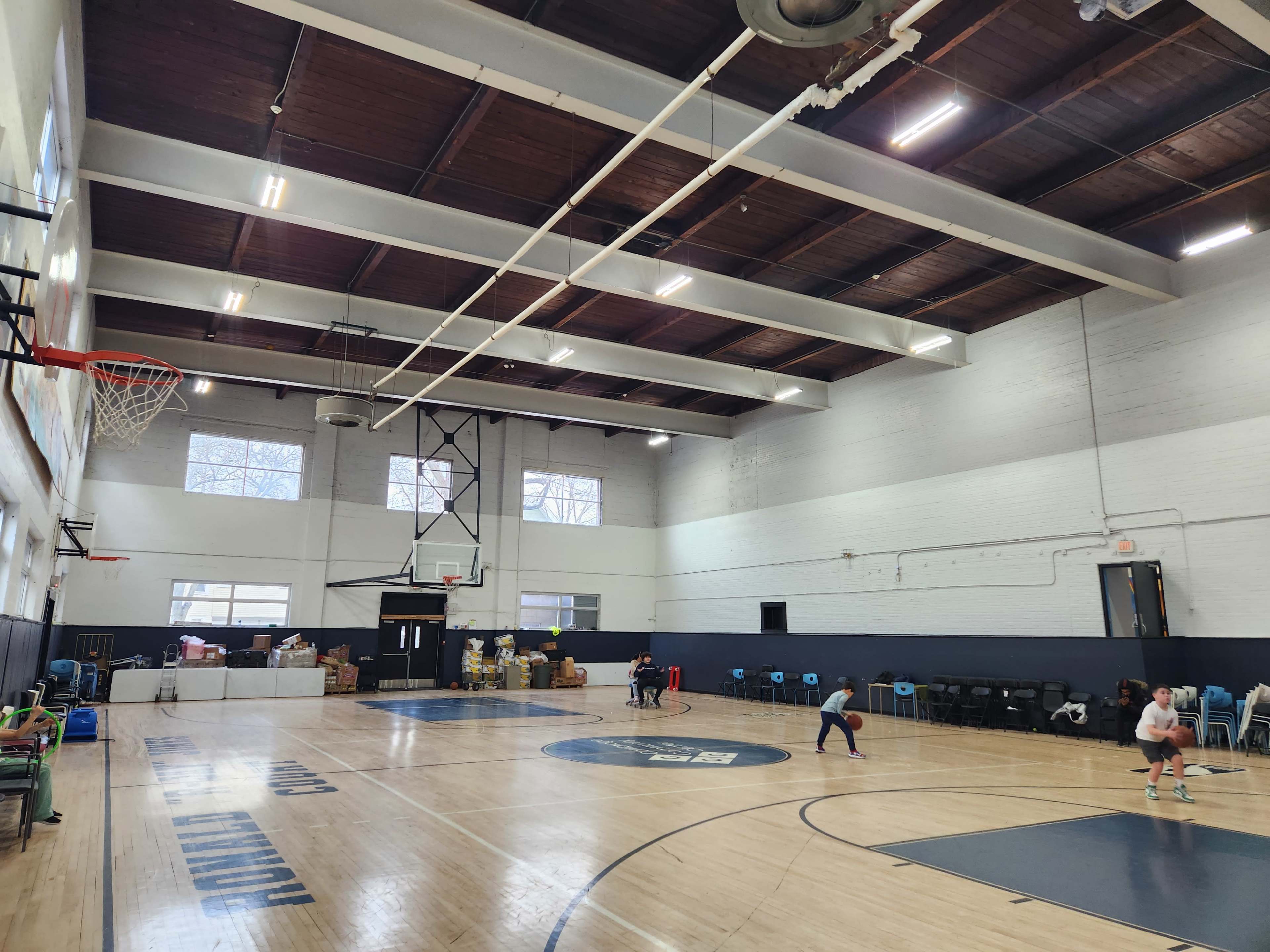A basketball court is shown with a high ceiling, featuring a hoop and two children playing while boxes and chairs are arranged along the walls.