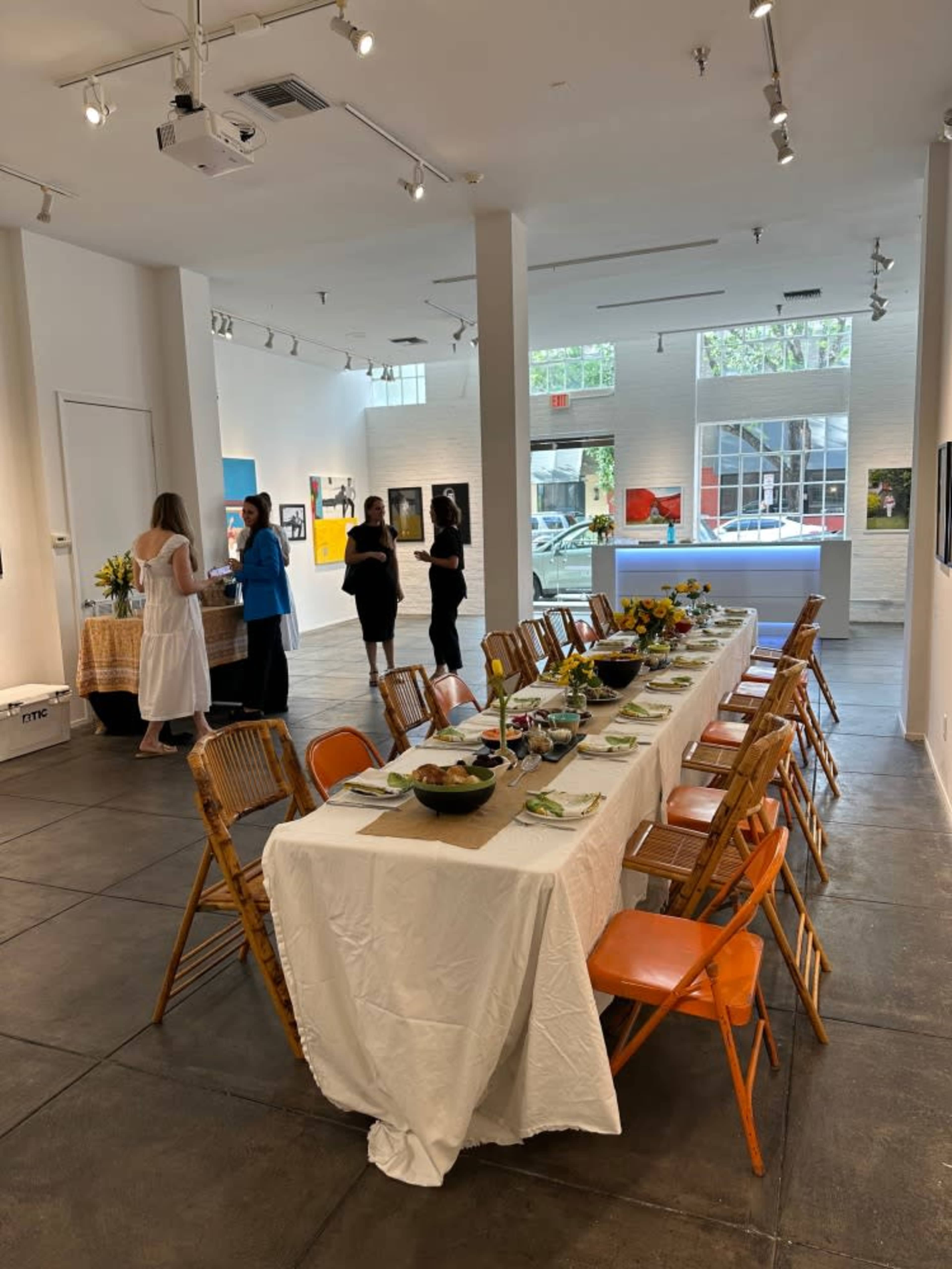 A long dining table is set in an art gallery, with guests mingling and a display of artworks on the walls.