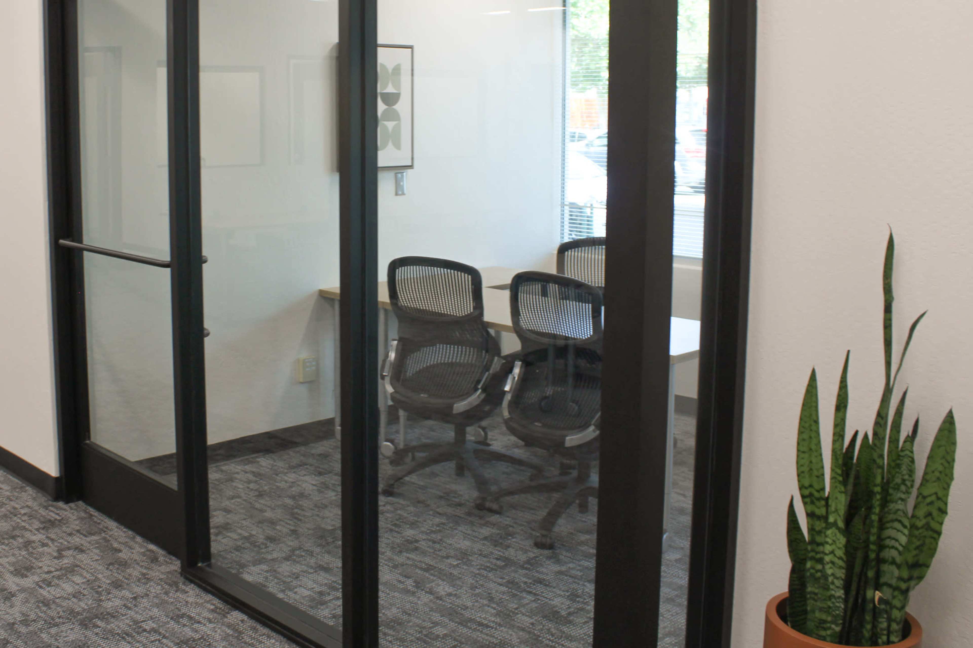 A glass-walled conference room featuring two office chairs and a small table is visible from a hallway.