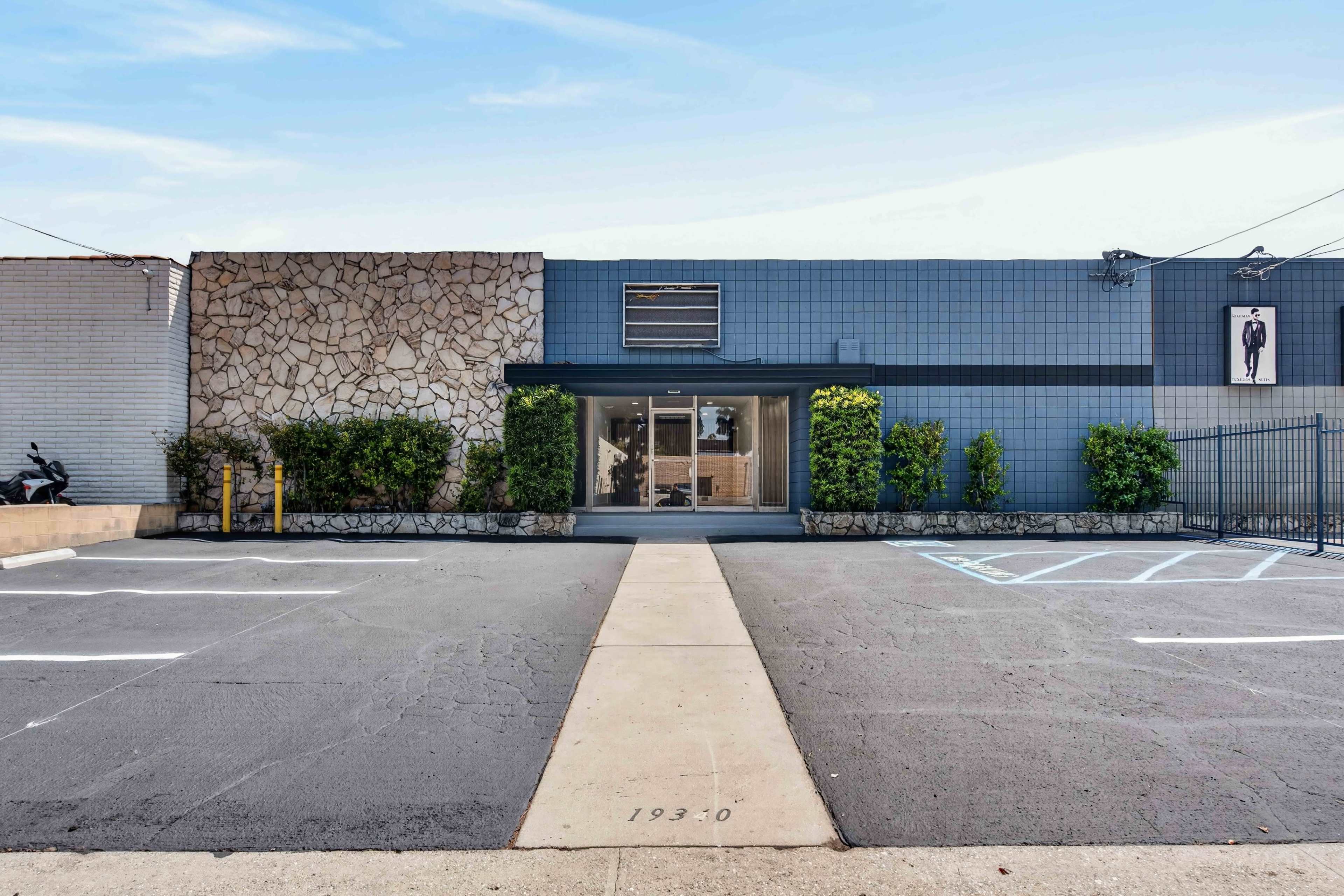 The image shows a modern one-story building with a stone and blue tile facade, featuring a central entrance and a parking lot in front.