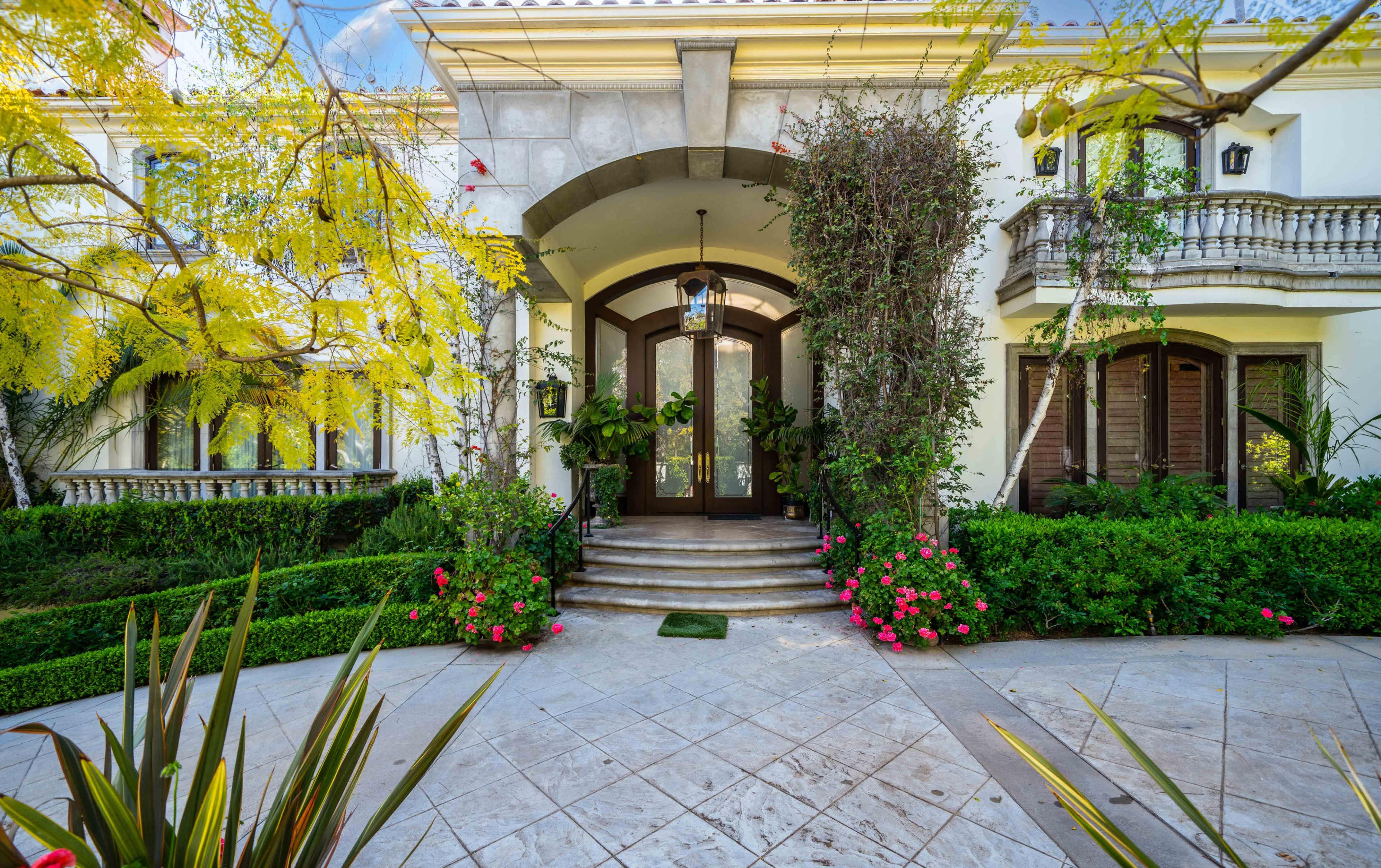 The image shows a grand entrance to a house, featuring arched wooden doors flanked by greenery and colorful flowers, with a stone pathway leading up to it.