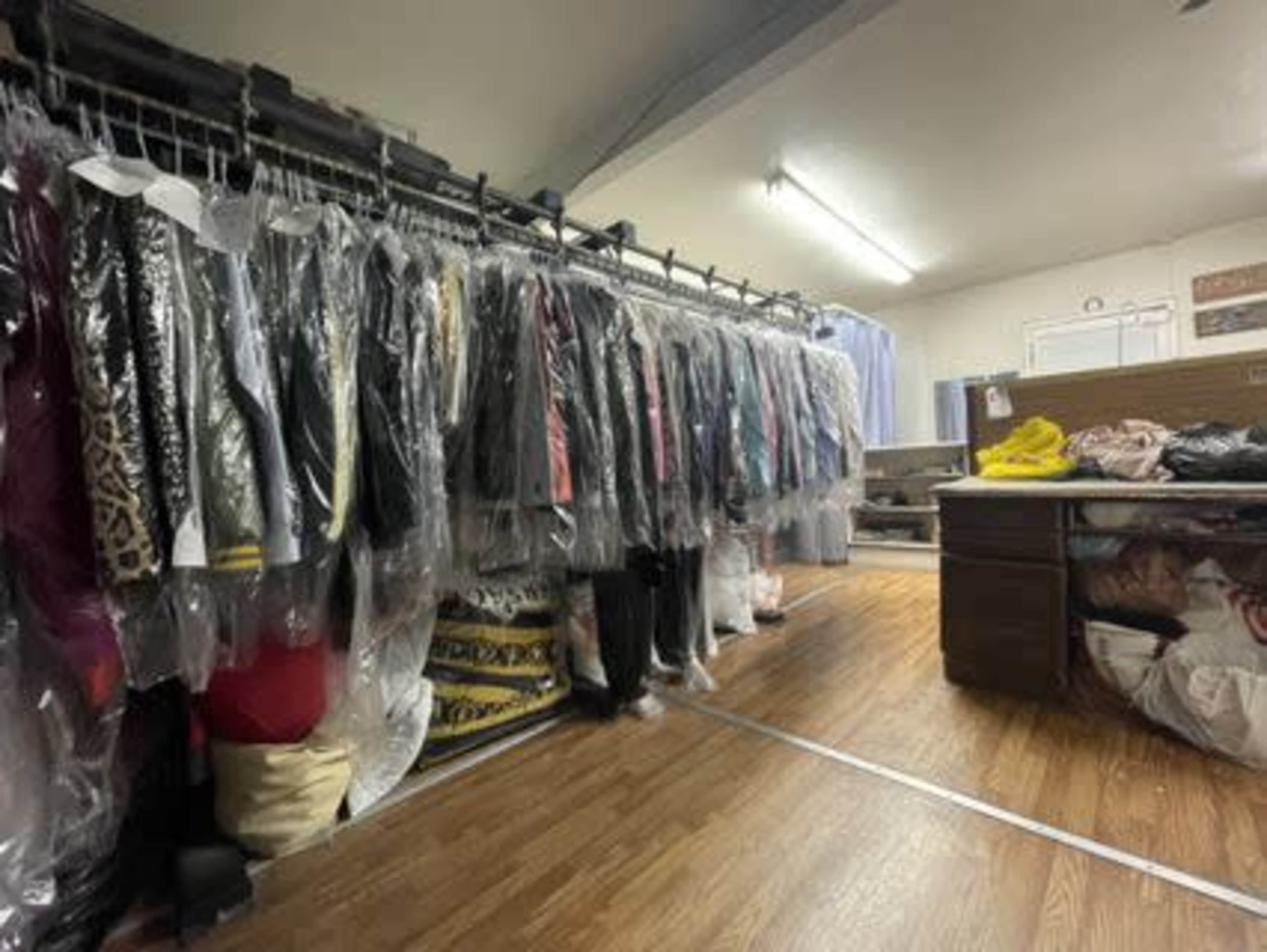 The image shows a row of garment bags hanging from a rack in a room with wooden flooring and various clothing items on a table in the background.