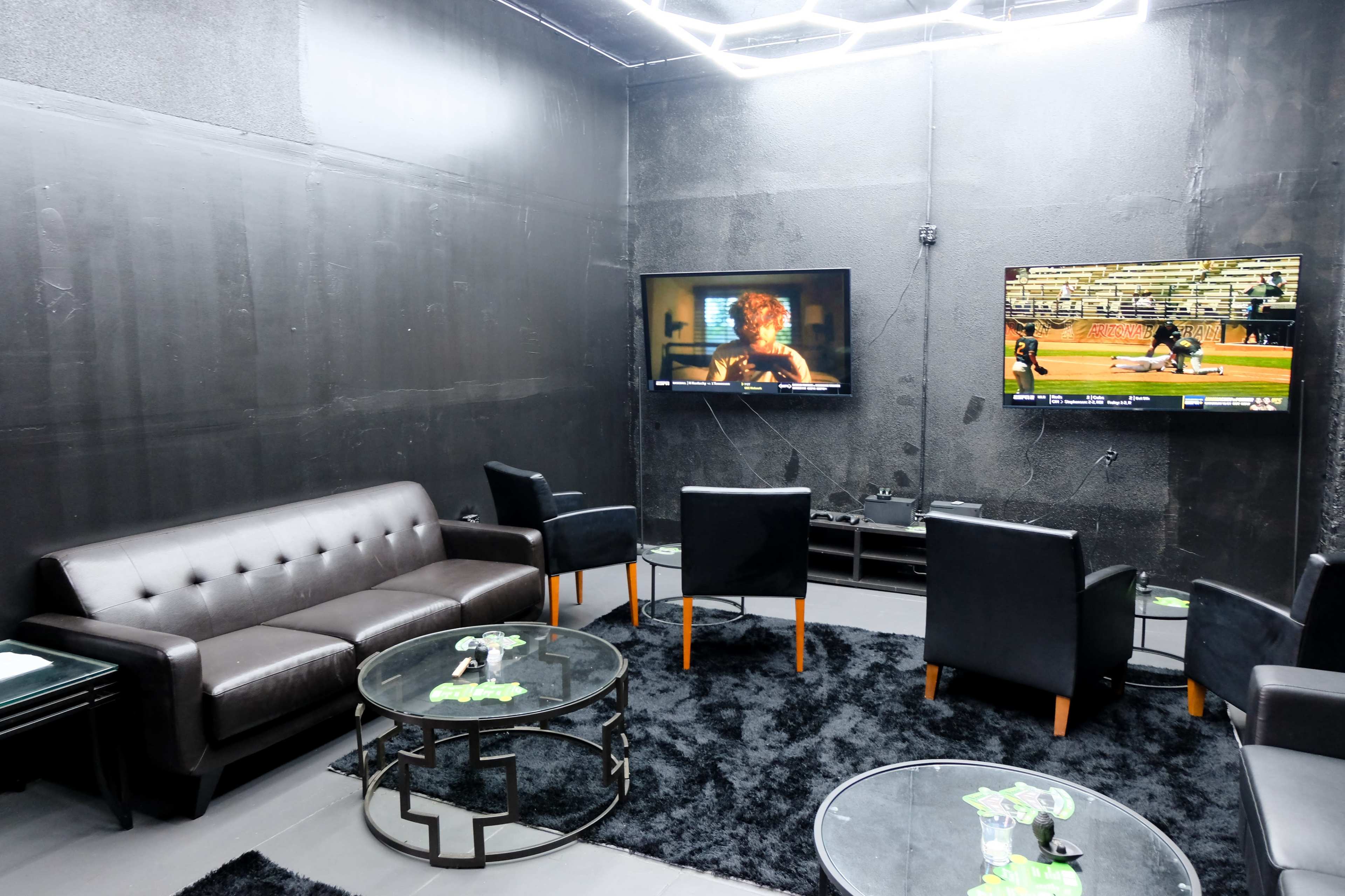 A dimly lit lounge area with black walls, a black sofa, several black chairs, and two televisions displaying different programs.