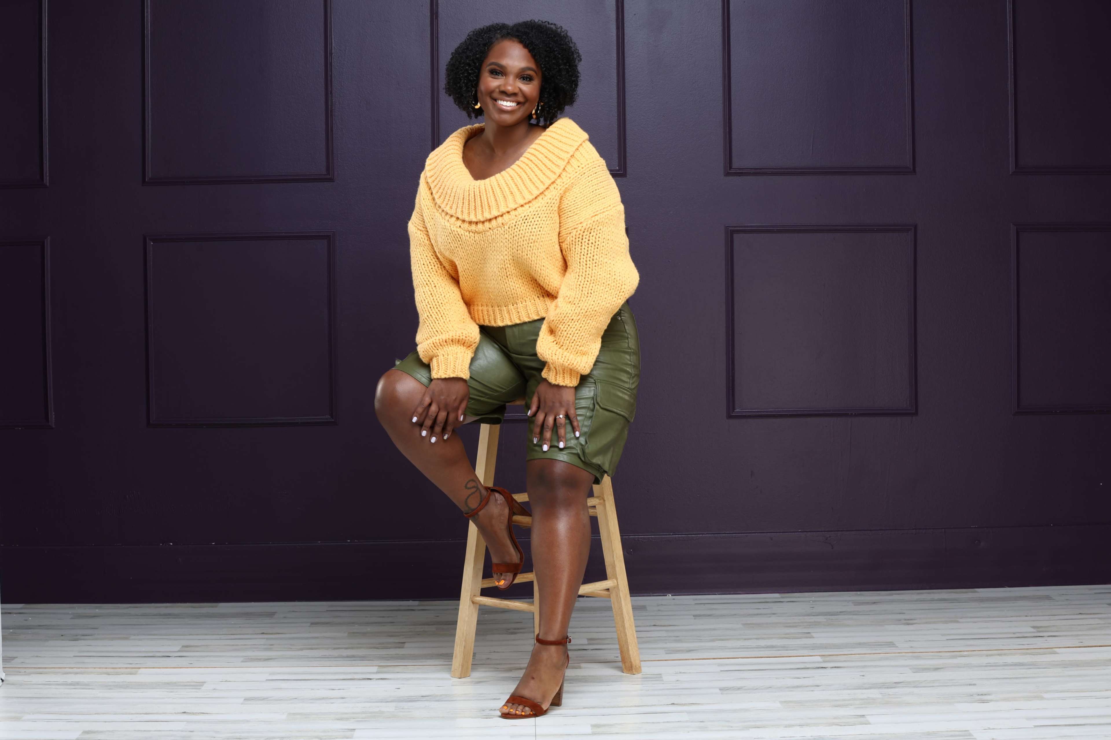 A woman sits on a wooden stool, wearing a yellow knitted sweater and green leather shorts, against a dark purple wall.