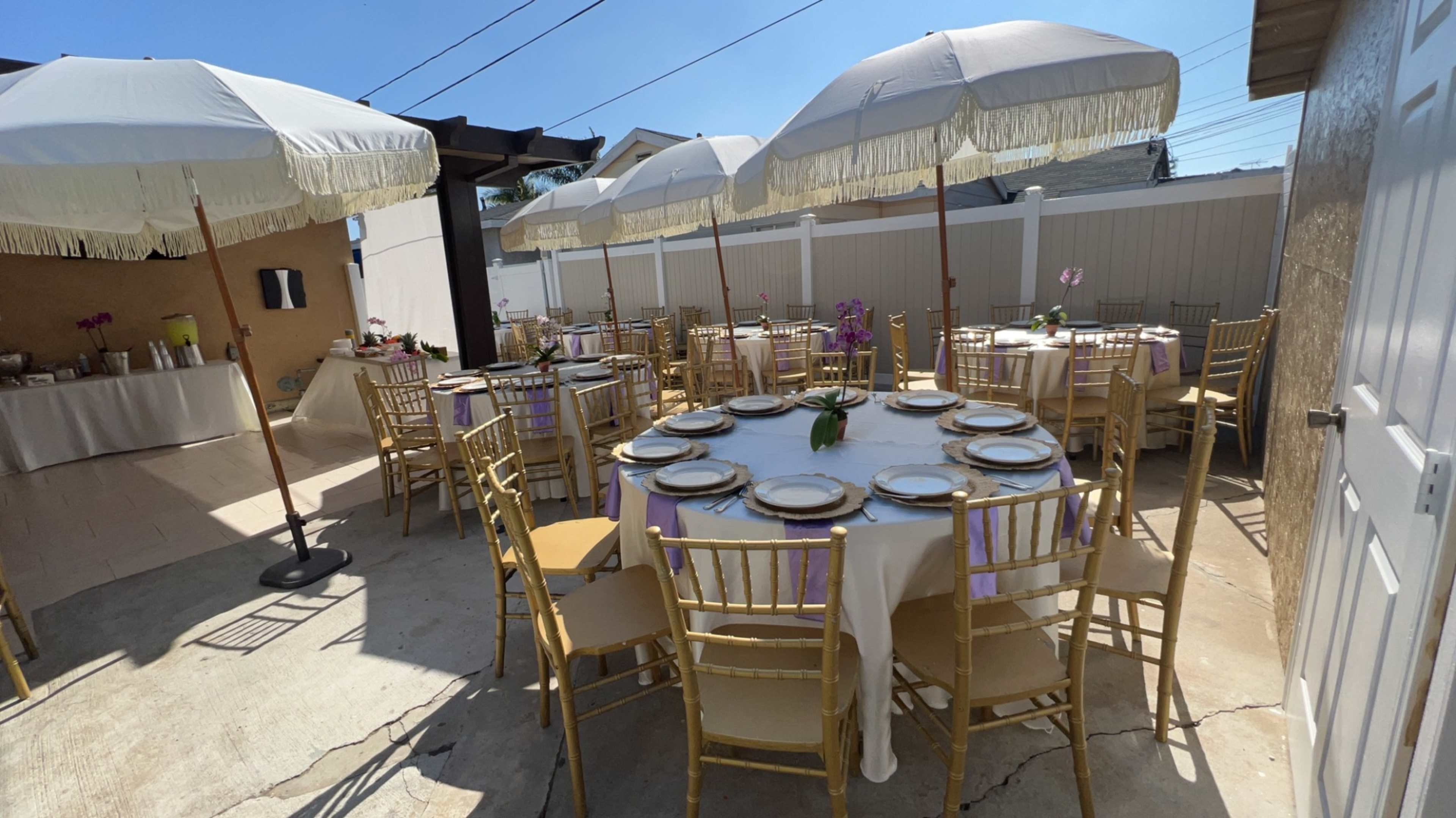 The image shows a decorated outdoor dining area with several round tables set with plates and surrounded by umbrellas for shade.