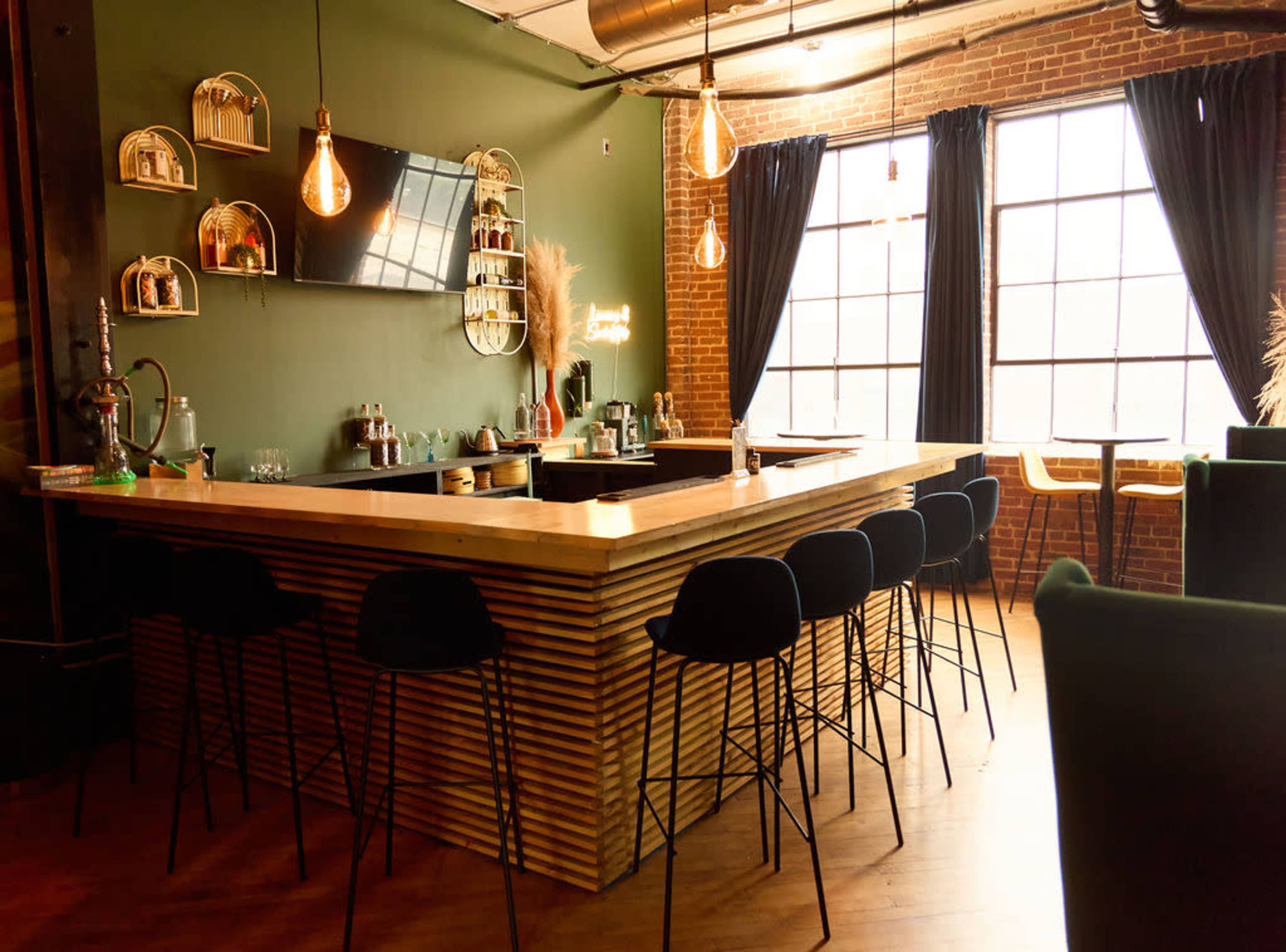 A modern bar area features a wooden counter, black stools, and large windows framed by dark curtains in a space with exposed brick walls.