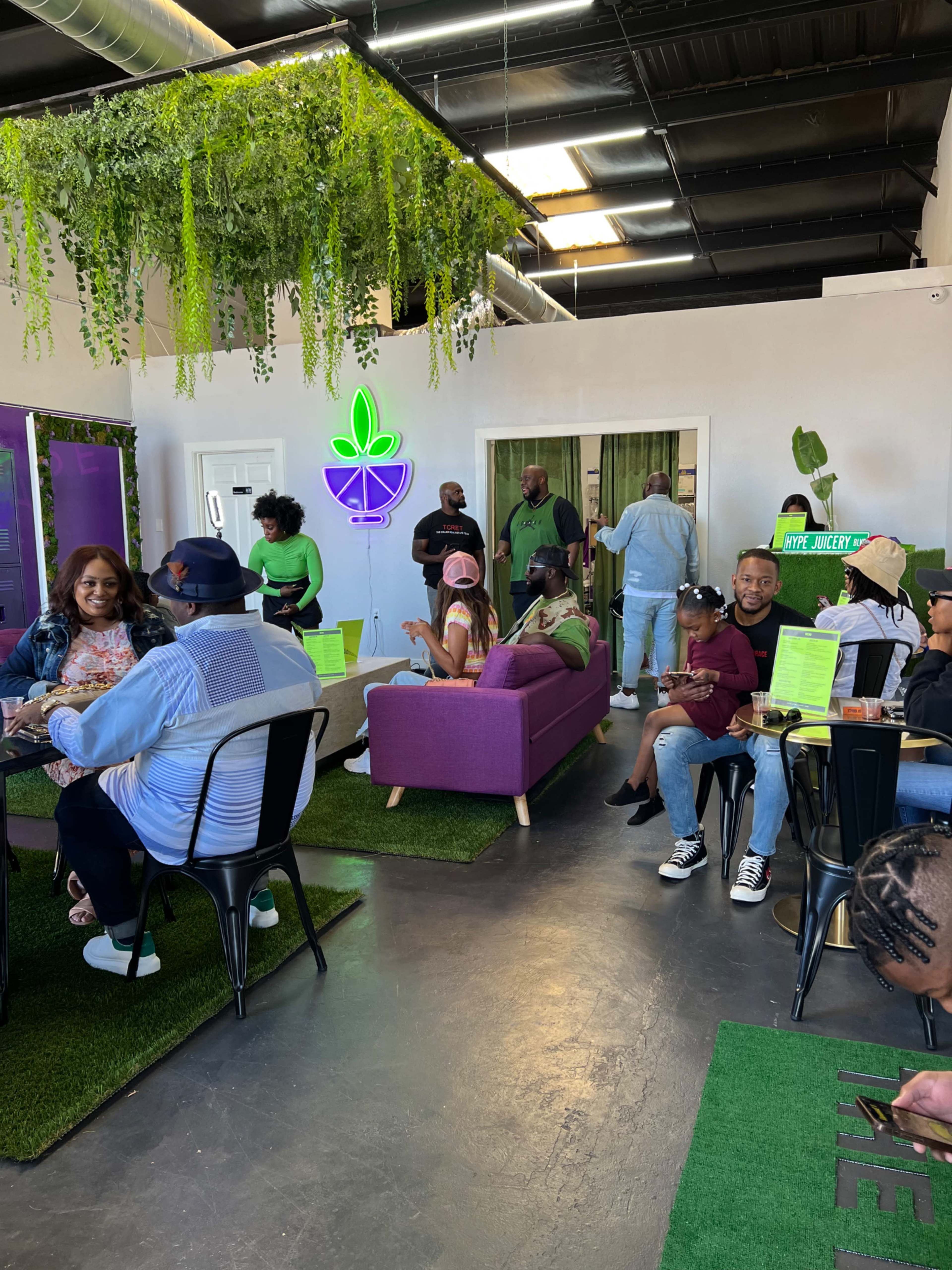 A vibrant indoor space features a green-themed decor with plants, seating areas, and people engaging in conversation and dining.