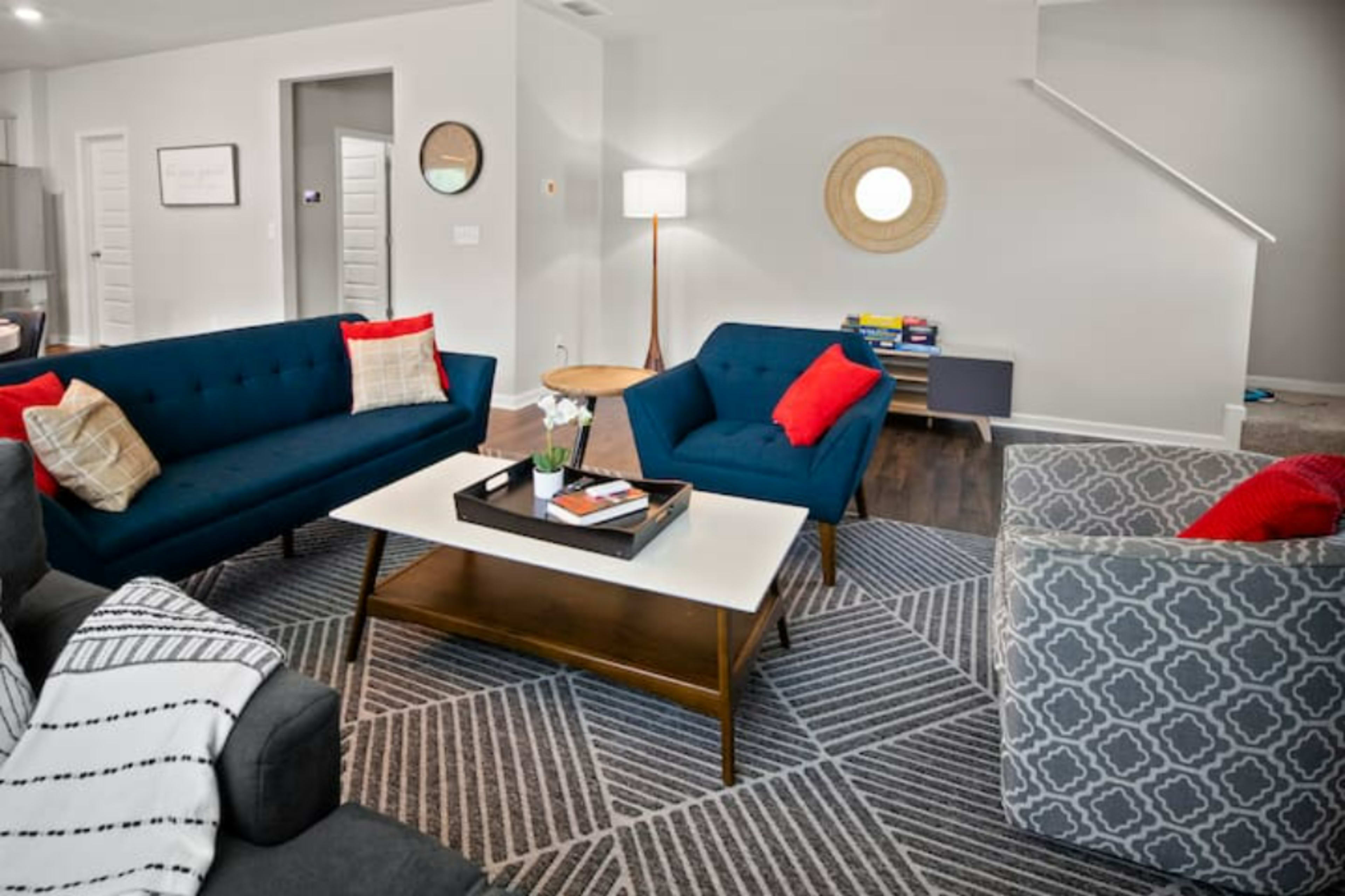 The living room features two teal sofas, a patterned rug, a coffee table with books and a plant, and a staircase visible in the background.