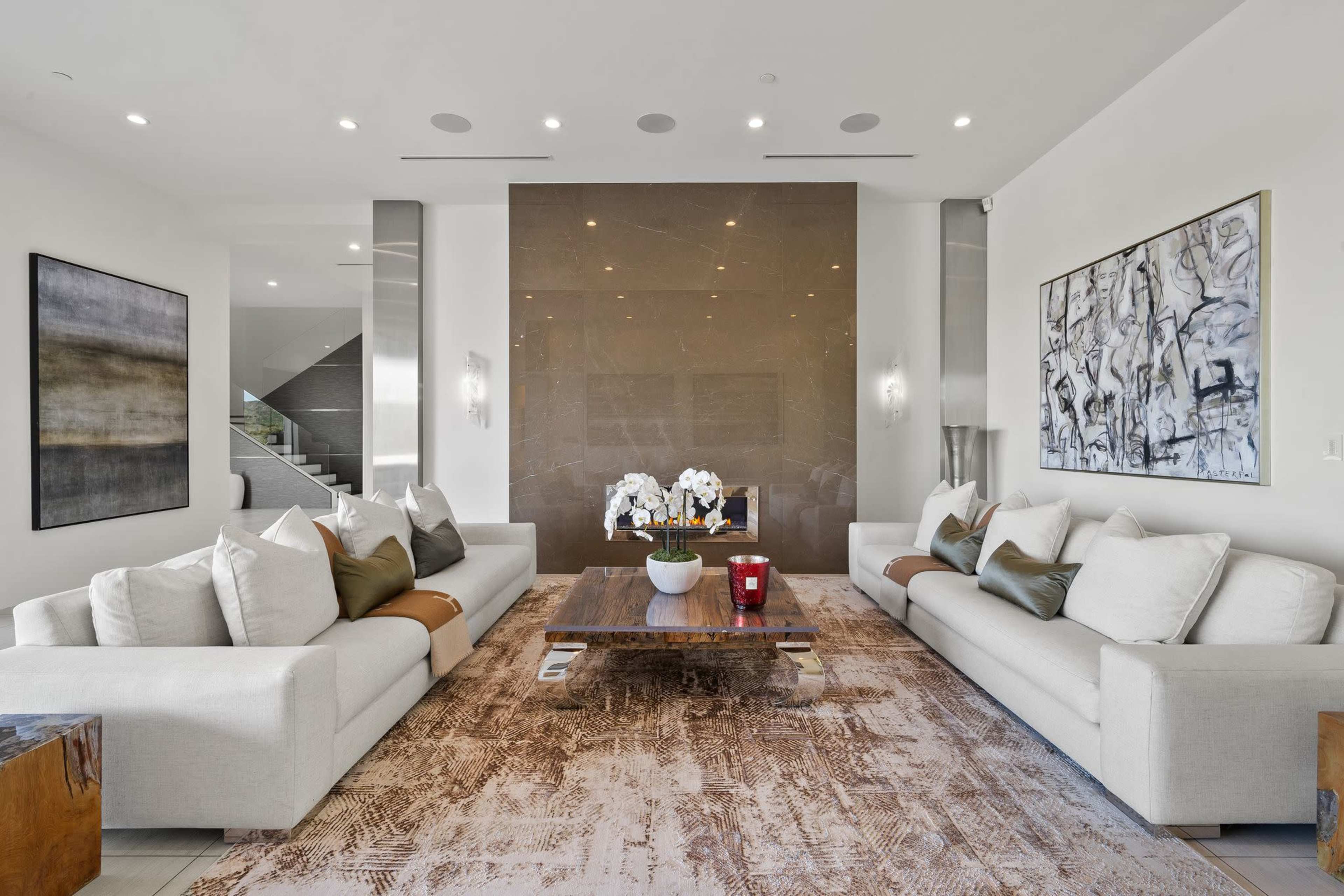 A modern living room featuring two light-colored sofas with decorative cushions, a wooden coffee table adorned with flowers, and a large stone wall at the back.