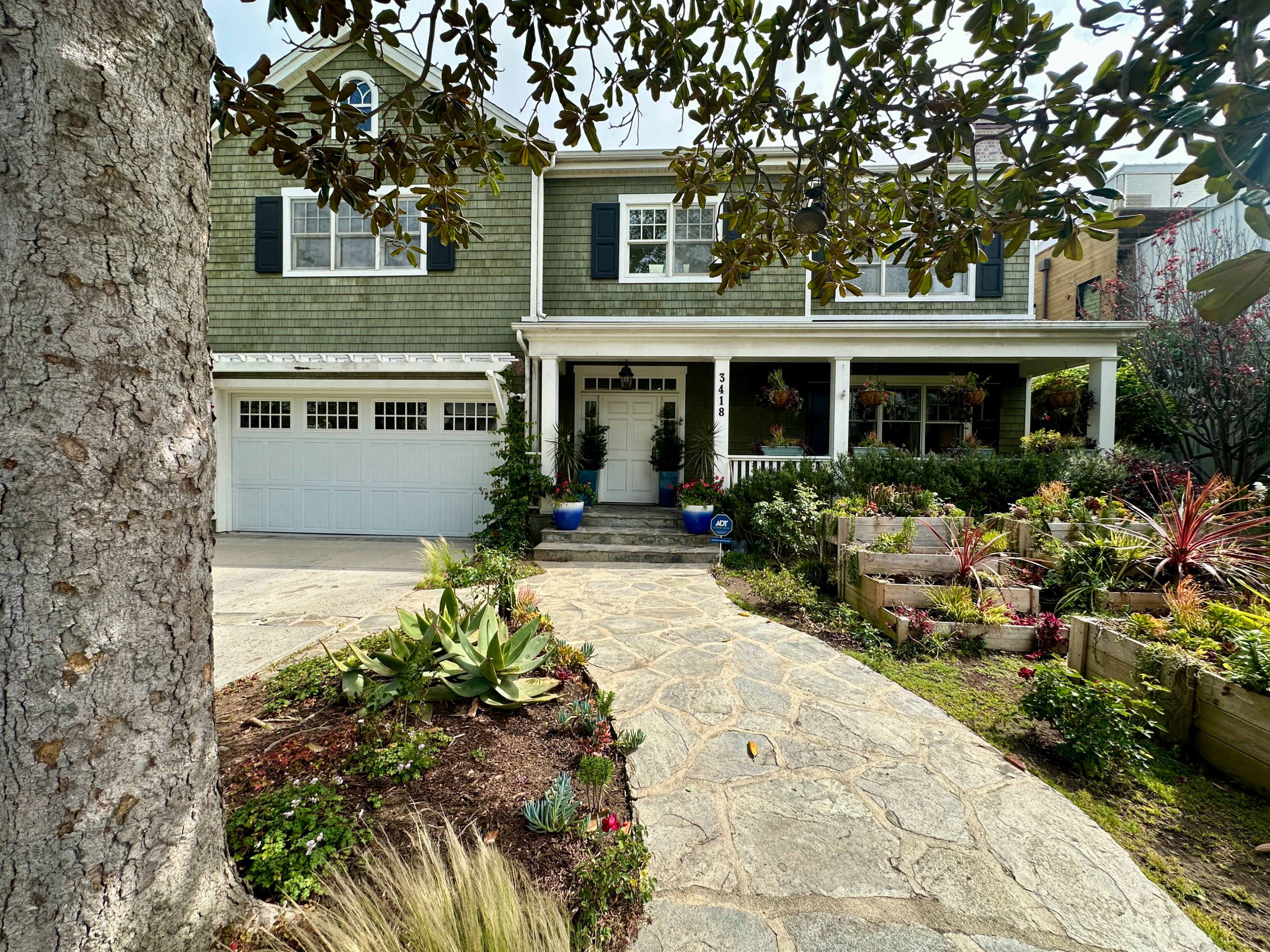 The image shows a two-story green house with white trim, a paved walkway, and a well-landscaped garden at the front.