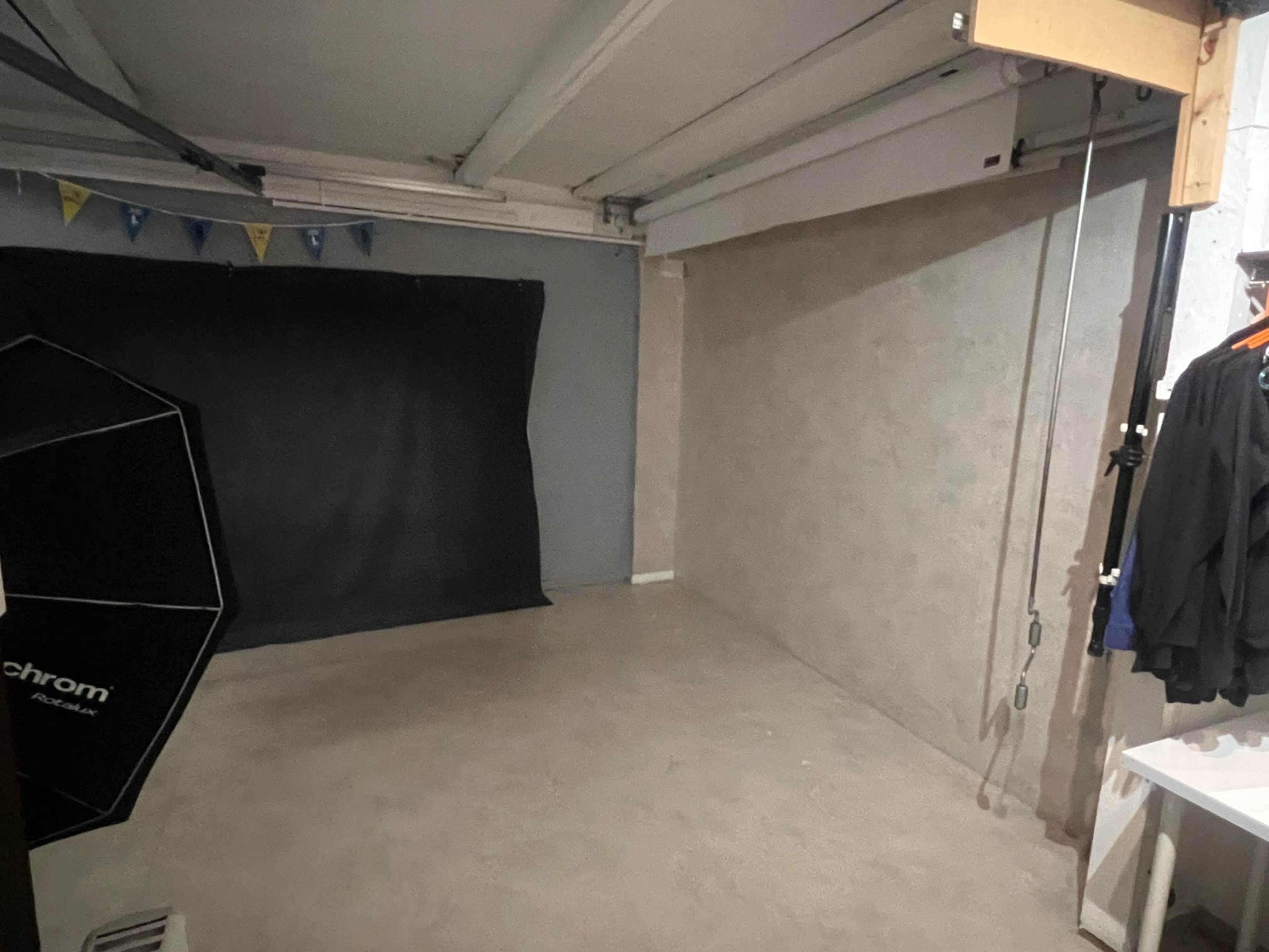 A spacious room with a gray floor and walls, featuring a black backdrop on one side, an umbrella light stand, and a few hanging garments.