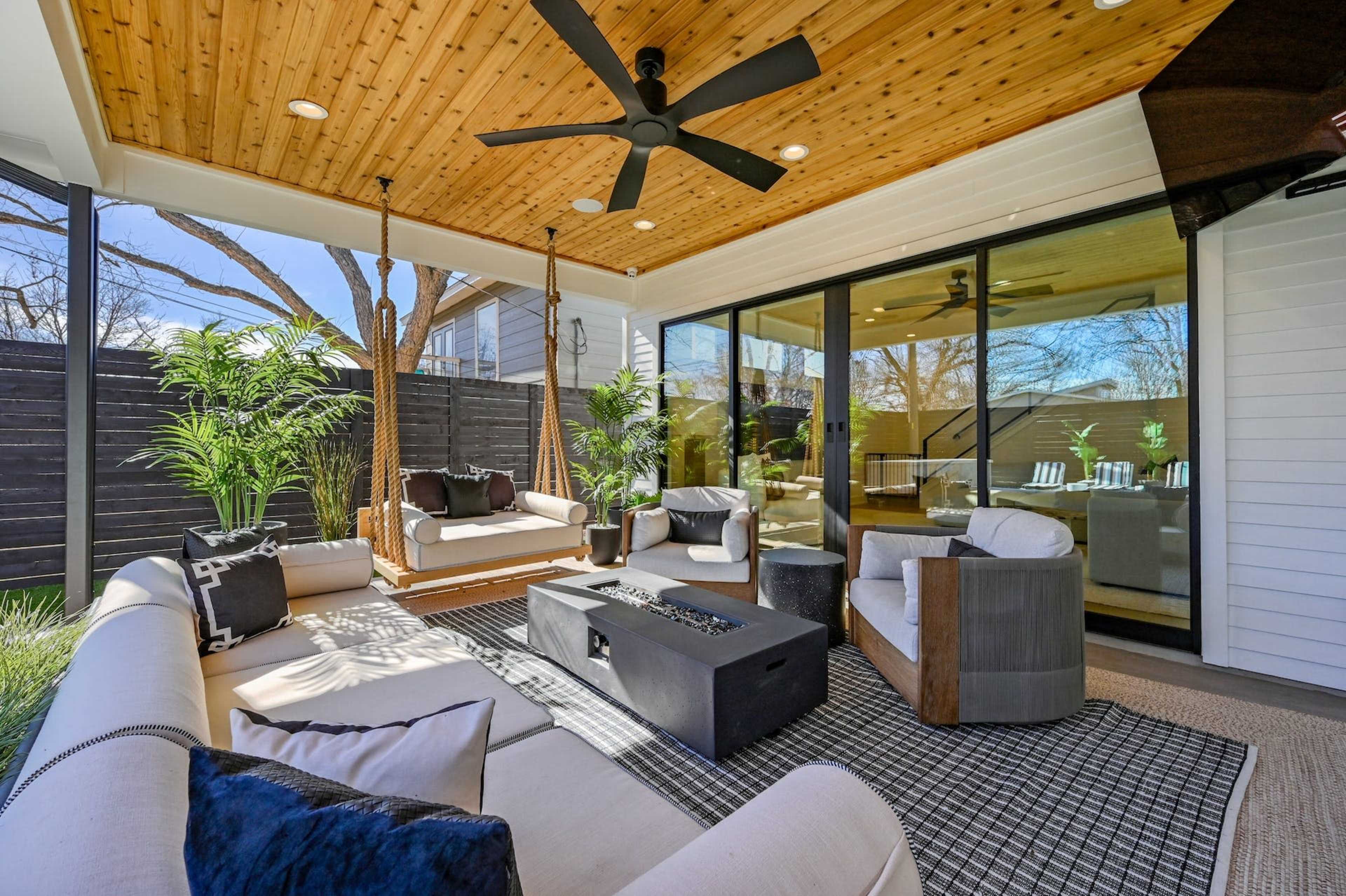 The image shows a modern outdoor seating area with a ceiling fan, comfortable couches, a coffee table with a fire pit, and greenery in a patio setting.