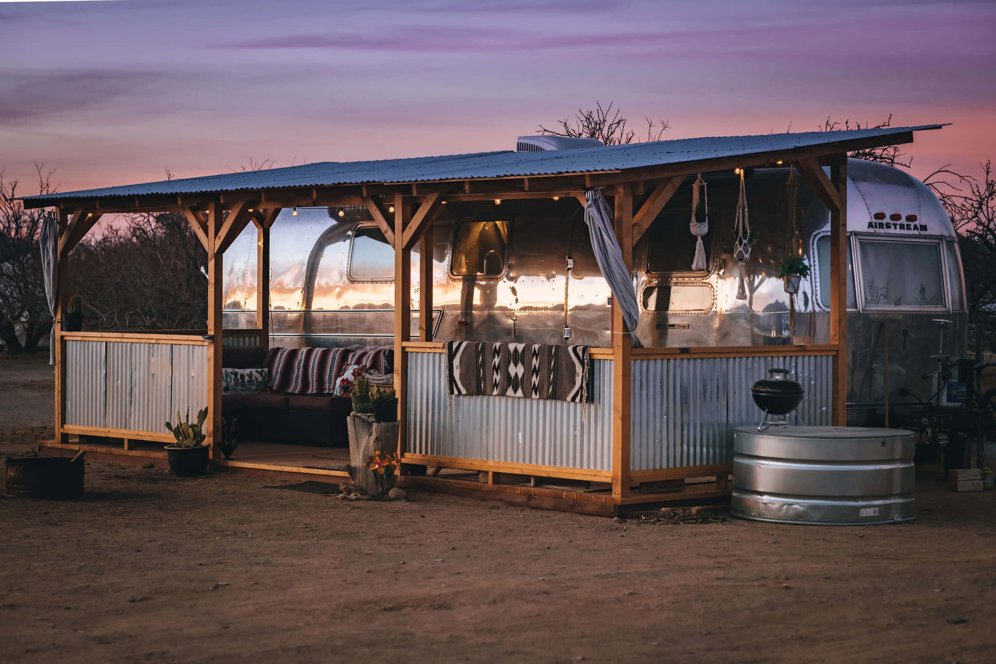 A silver Airstream trailer is set on a desert landscape, surrounded by a wooden porch with seating and decorative plants, as the sky displays hues of purple and orange at sunset.