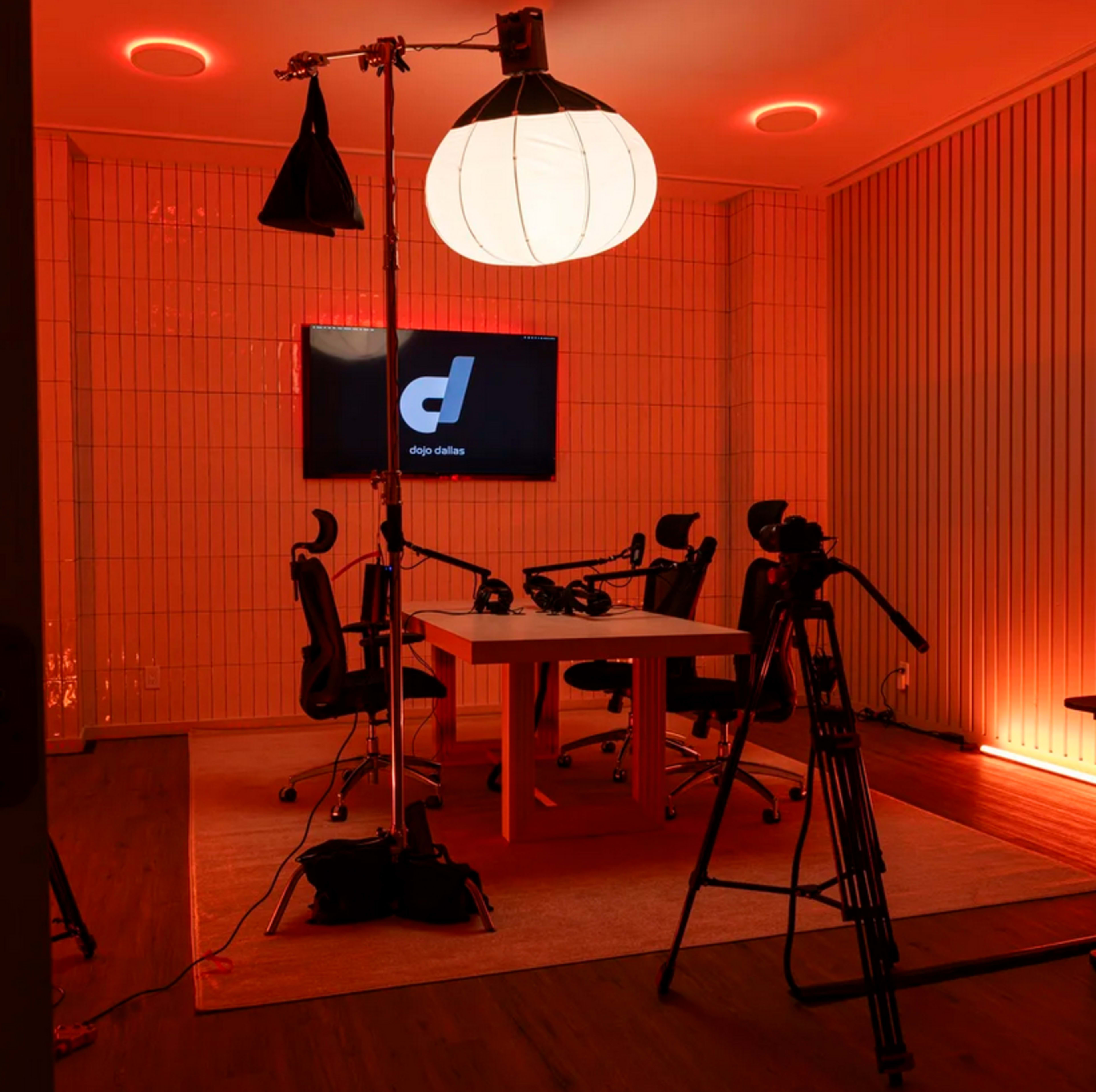 The image shows a podcast recording studio with a large round light fixture, a central table, and multiple cameras set up in a room illuminated with red lighting.